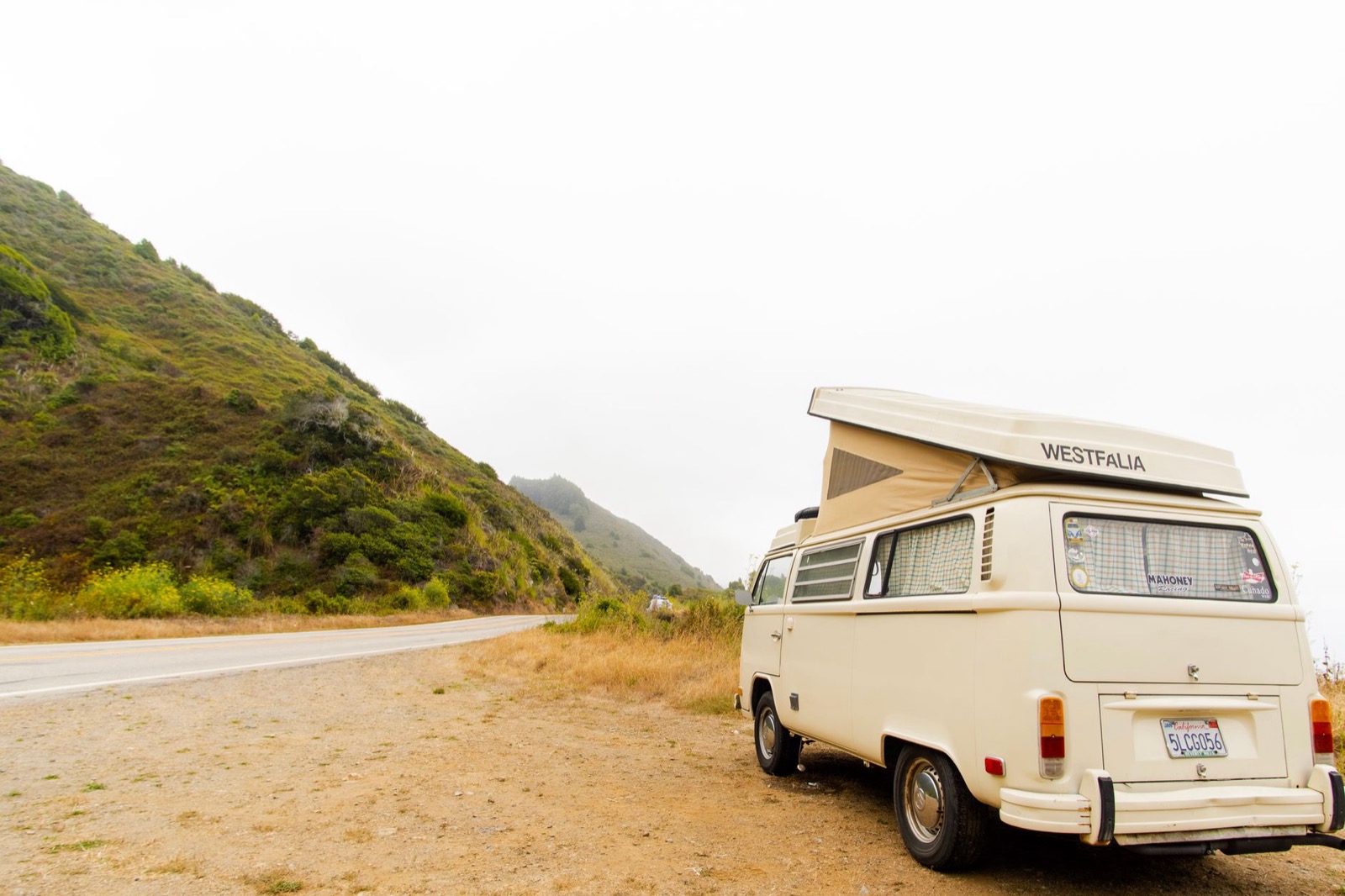 A pop top camper van parked on a gravel shoulder beside a coast road under low cloud