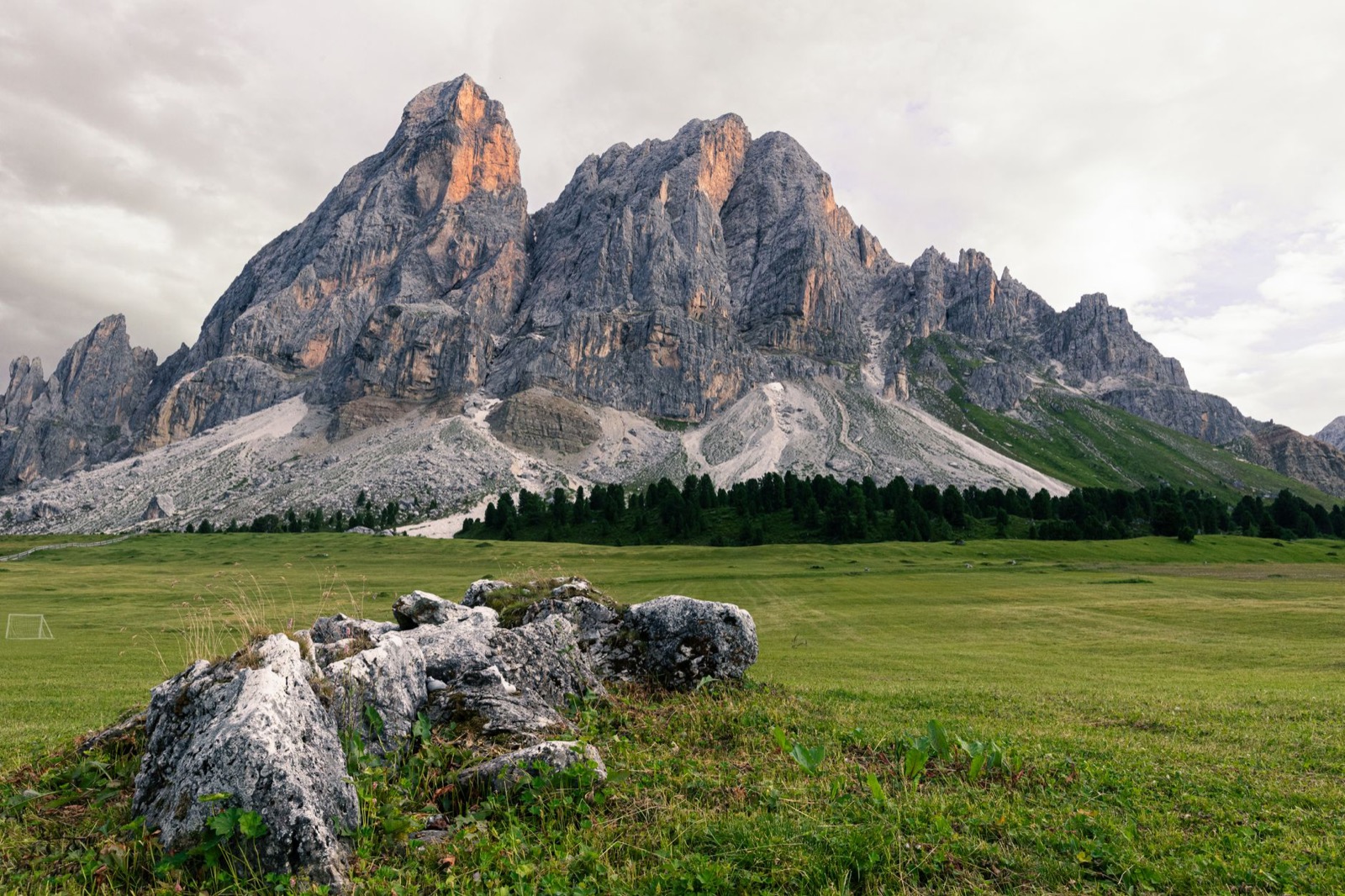 A jagged limestone peak above a green alpine meadow under a pale overcast sky