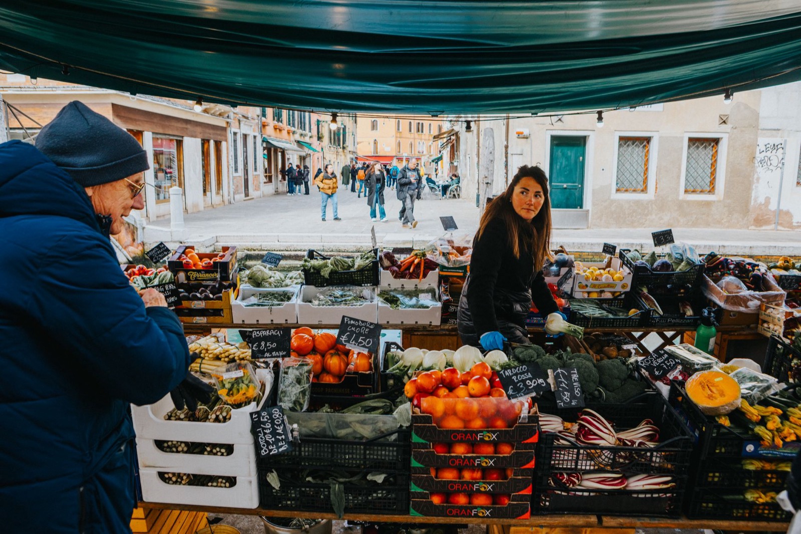 A vegetable stall under a green awning on an Italian street, crates of tomatoes and radicchio with chalked euro prices