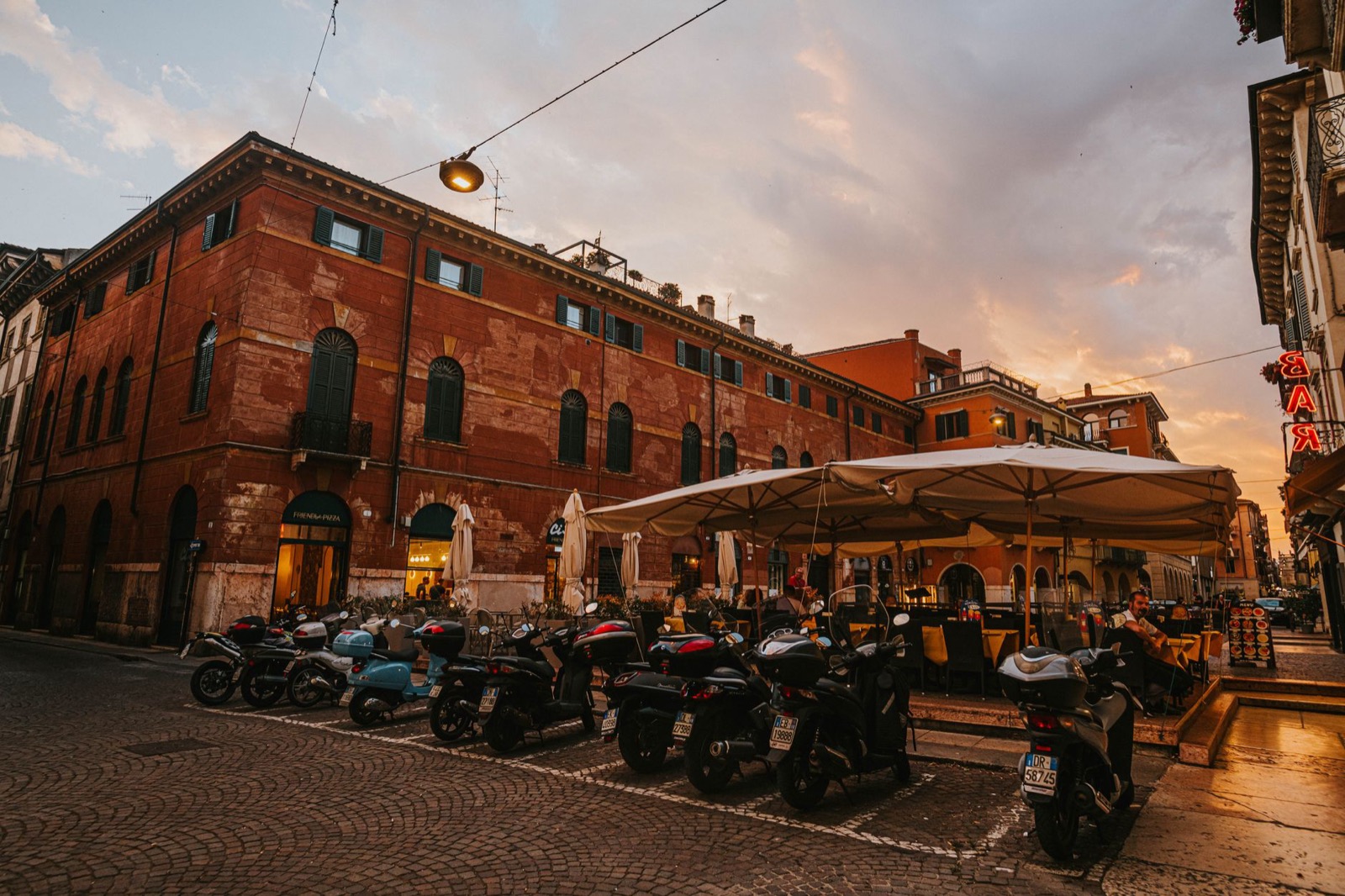 Scooters parked in a row across an Italian square at sunset, a cafe terrace under a white awning behind them