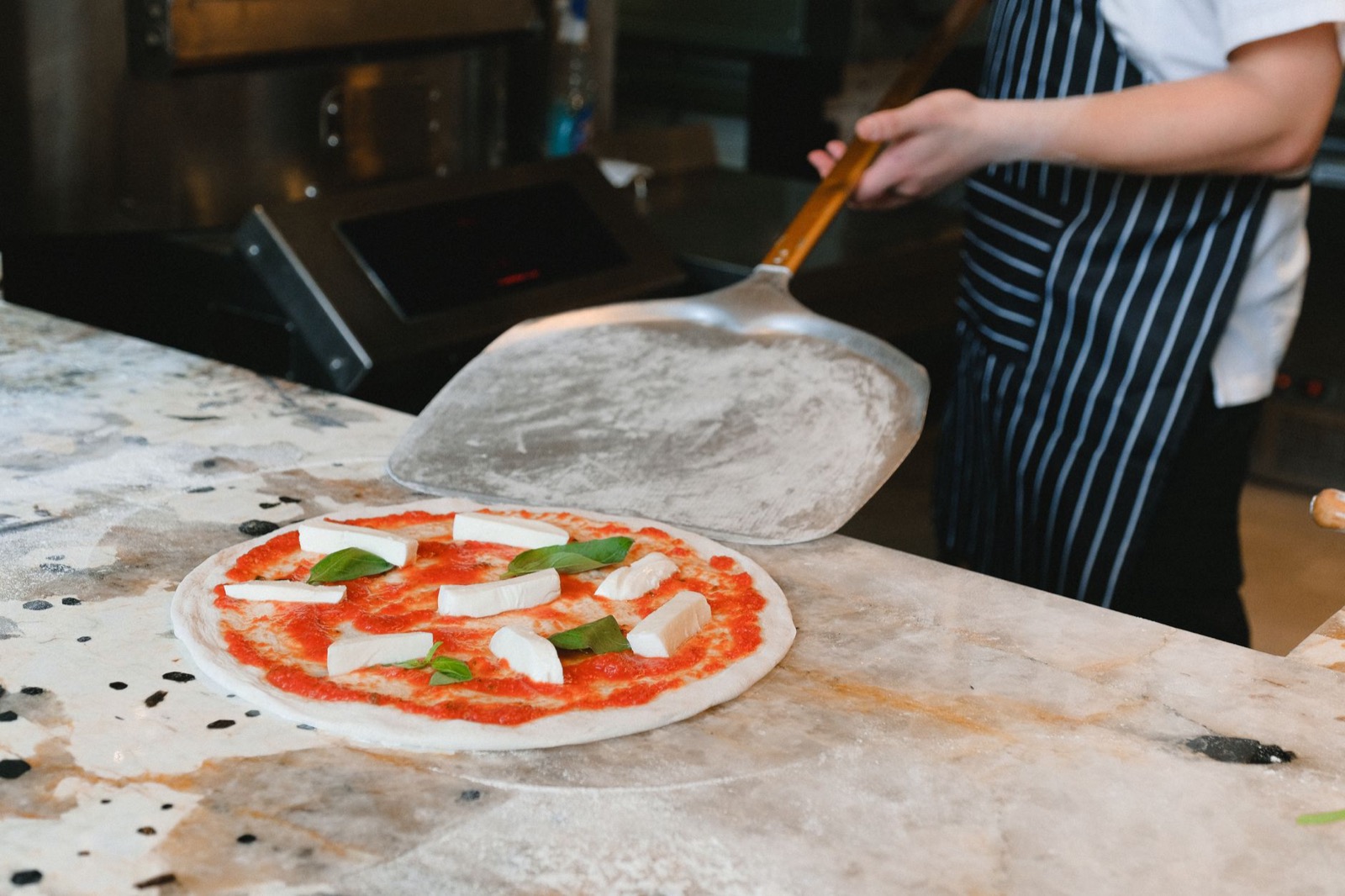 A raw margherita topped with mozzarella and basil on a floured marble counter, a pizzaiolo lifting the peel behind it