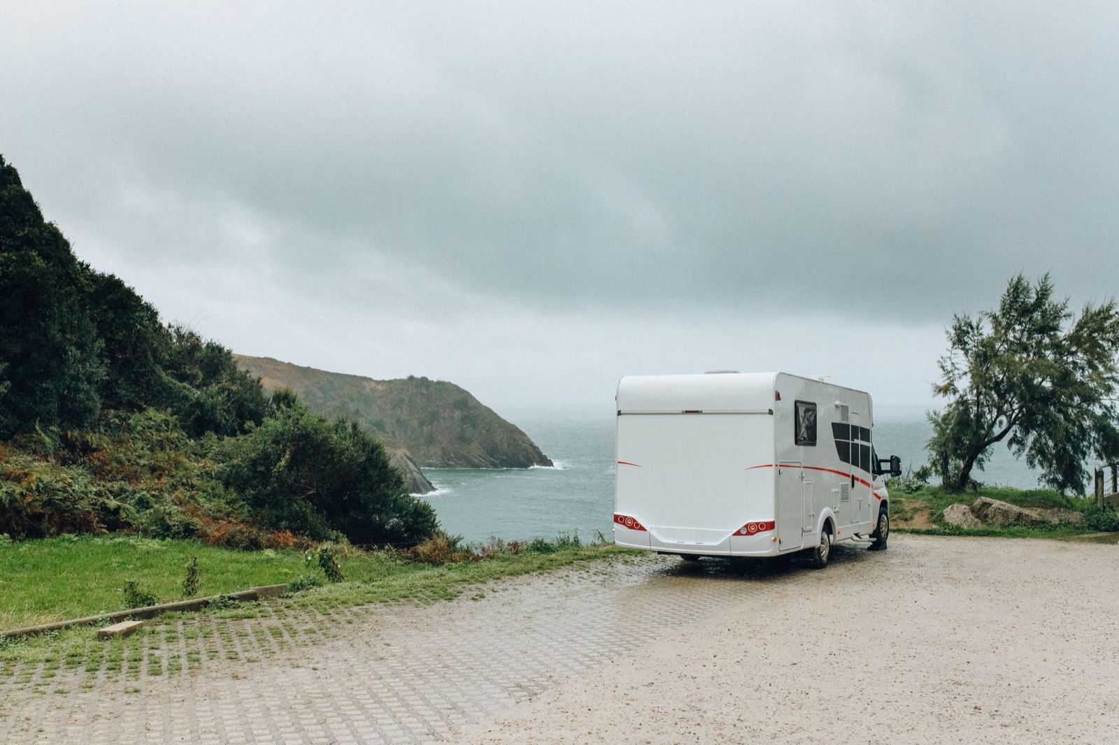 A white motorhome parked alone on a gravel area above a grey sea, a green headland beyond