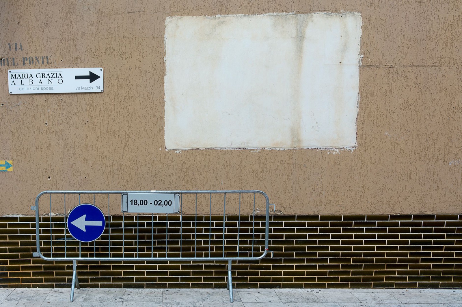 A metal barrier closing an Italian street, carrying a blue arrow sign and a plate reading 18,00 to 02,00