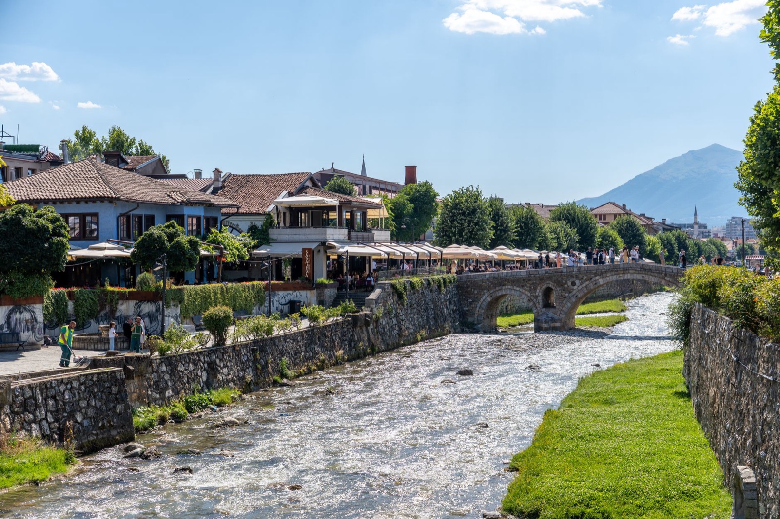 A stone arched bridge and riverside cafes under white umbrellas along a shallow river in an old town