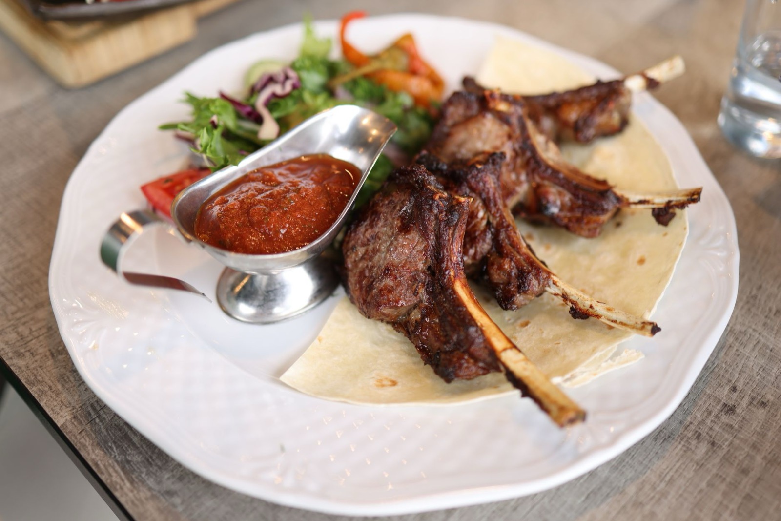 Grilled lamb chops on a flatbread with a side salad and a jug of red sauce