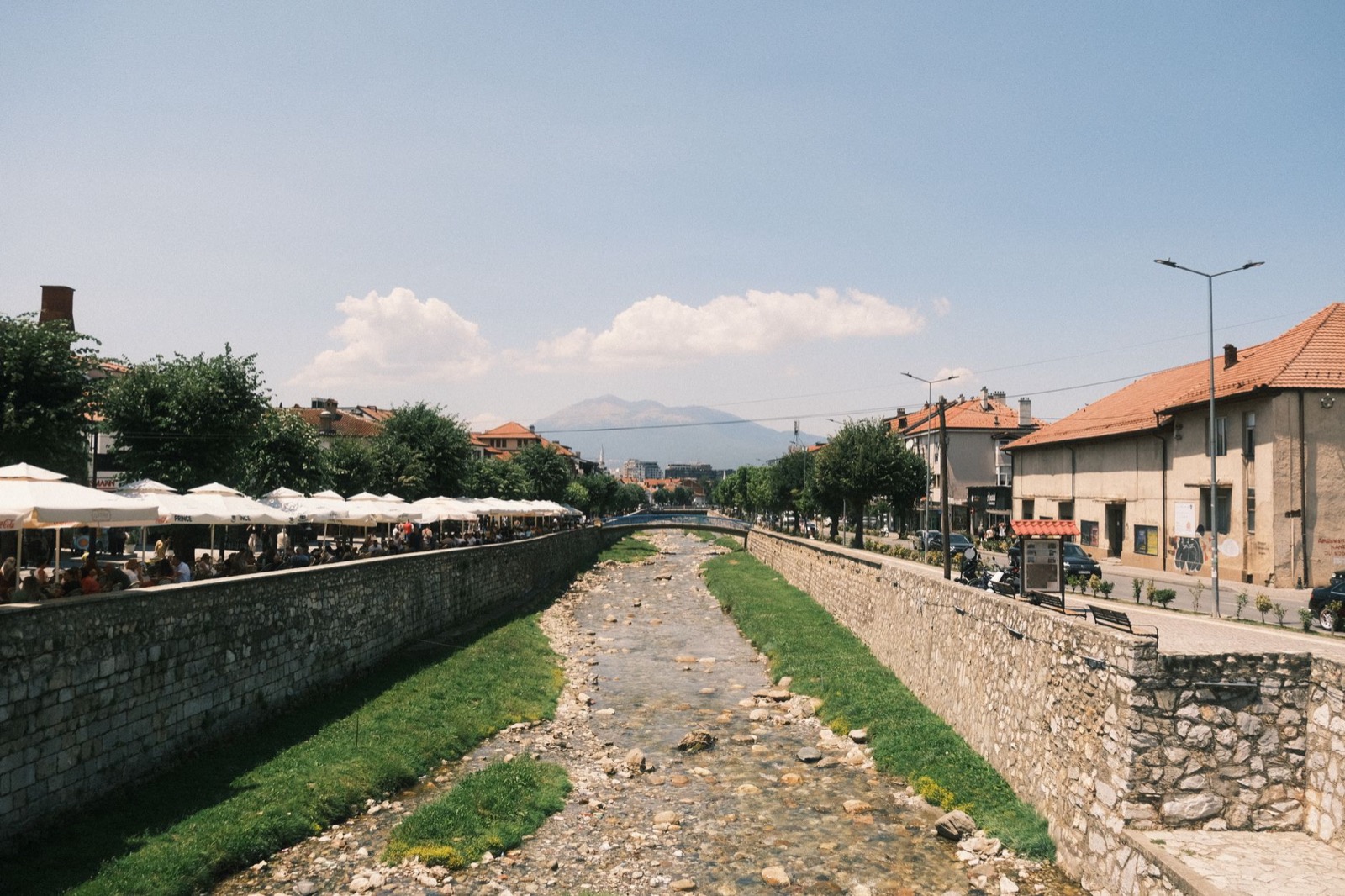 A shallow river between stone embankments through a town, with rows of cafe umbrellas along one bank