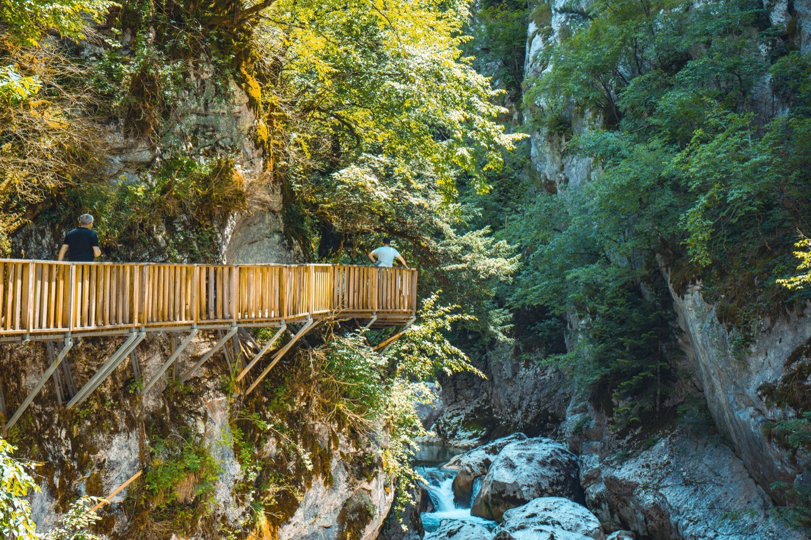 A wooden walkway pinned to a cliff face above a rocky stream in a wooded gorge