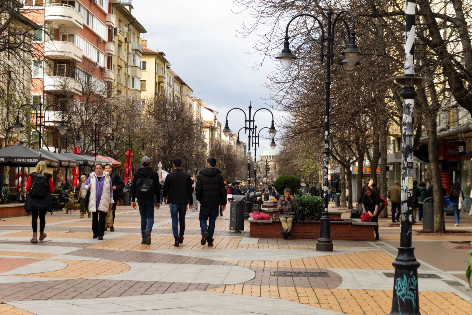 A broad pedestrian boulevard with ornate lamp posts, bare trees and patterned paving, busy with people