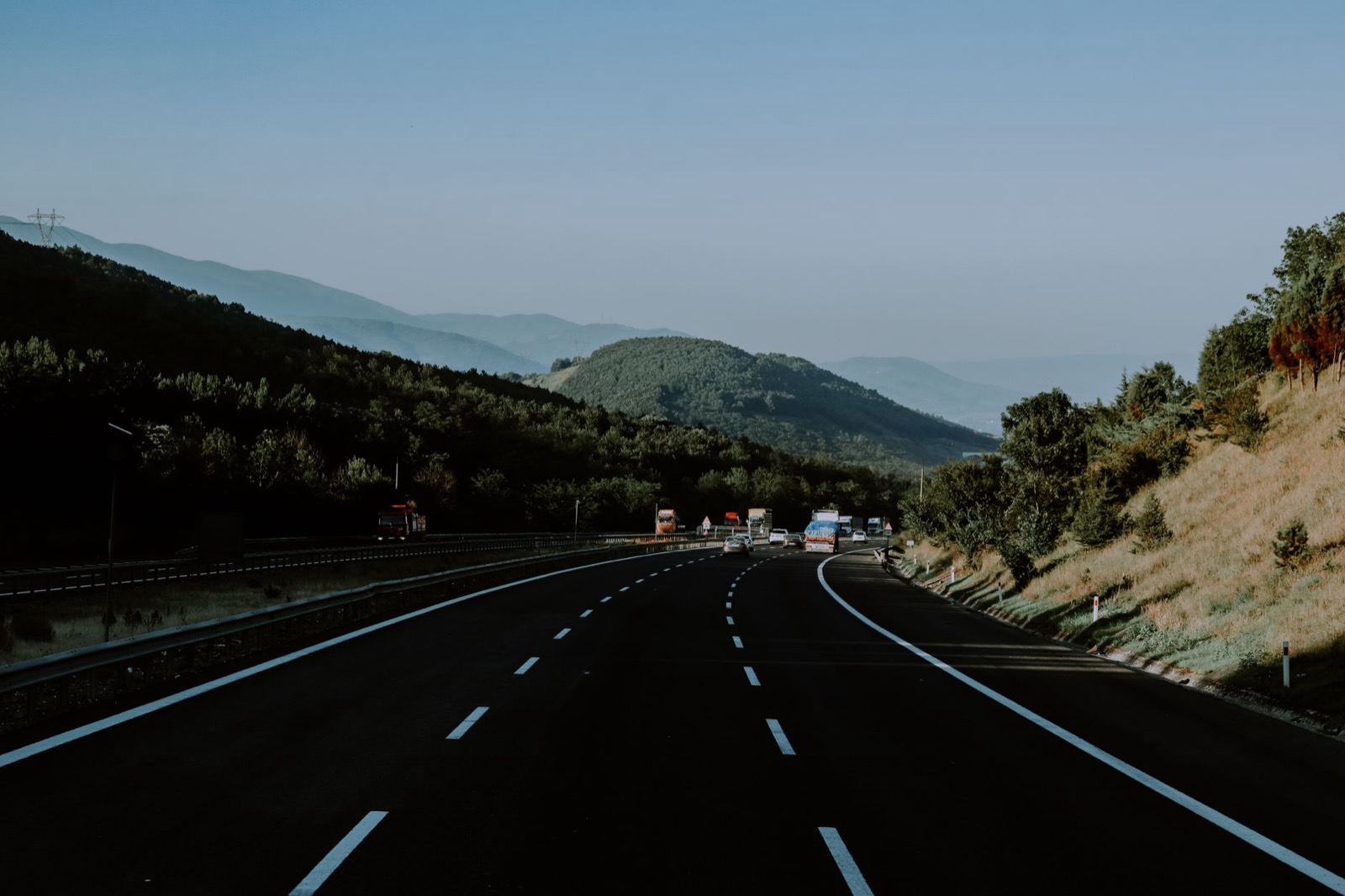 A new dual carriageway curving towards hazy forested hills with a few lorries ahead