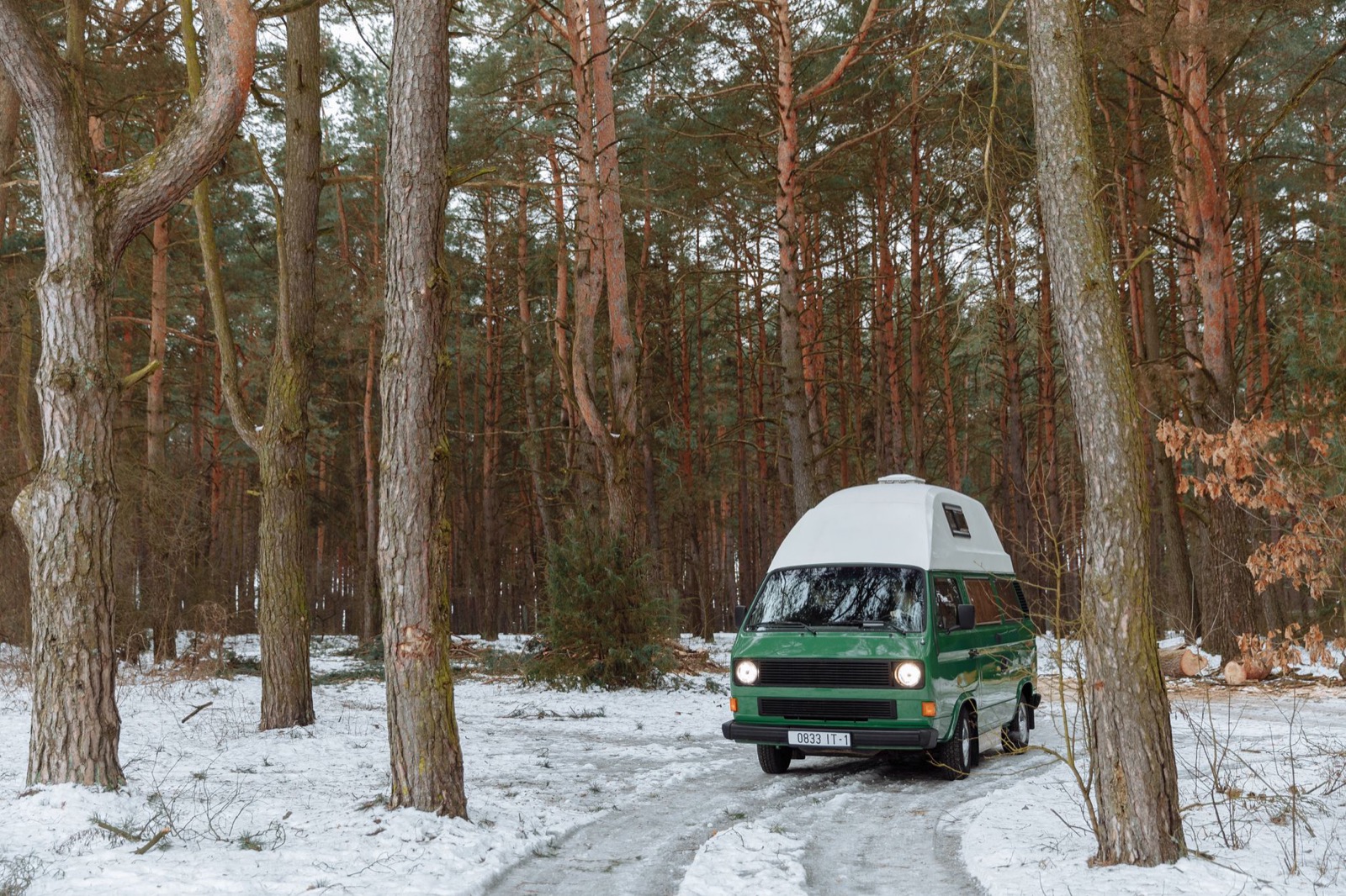 A green high-top camper van on a snowy track through pine forest, headlights on