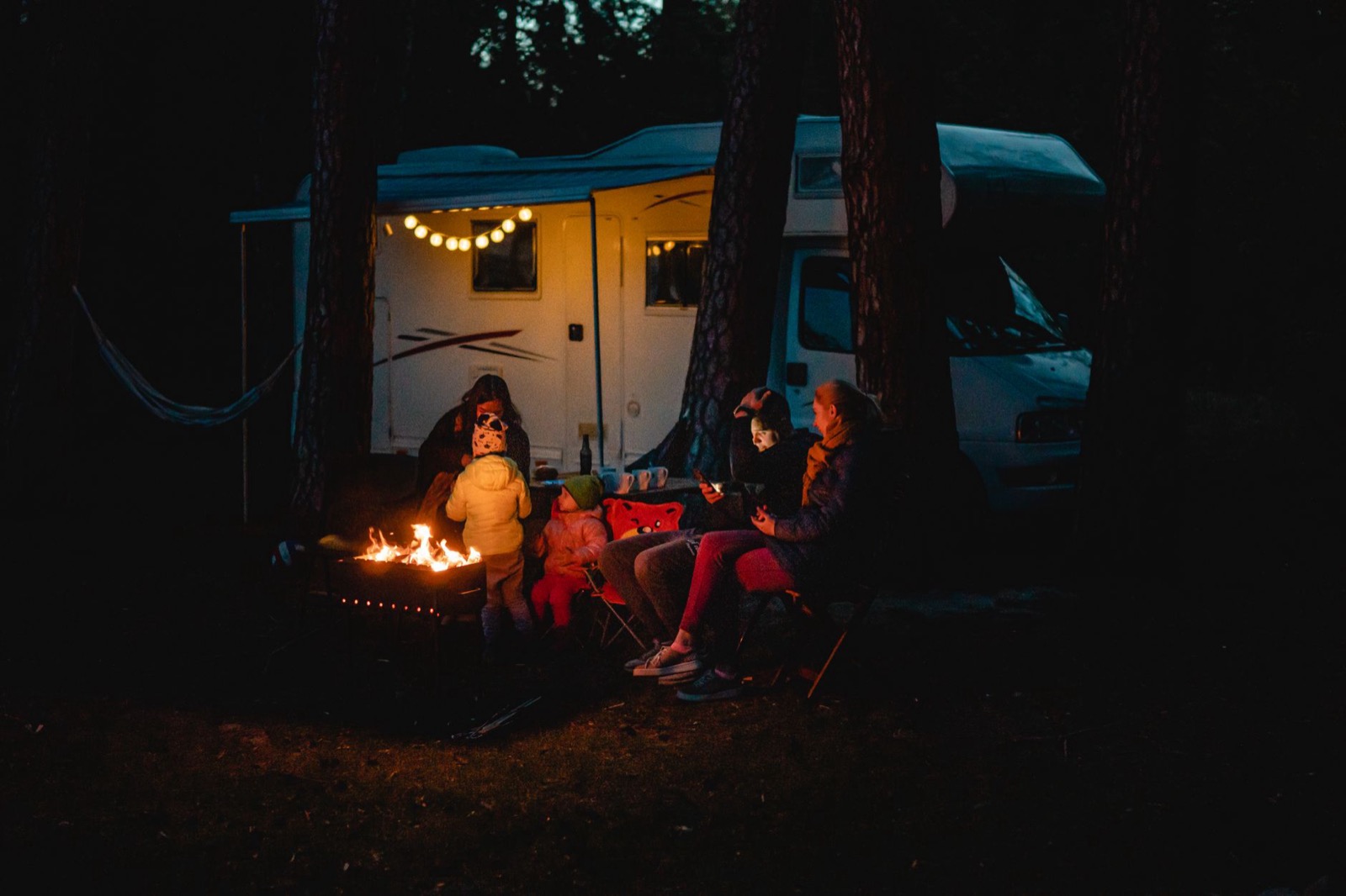 A family sitting round a campfire beside a lit motorhome in dark woods at night