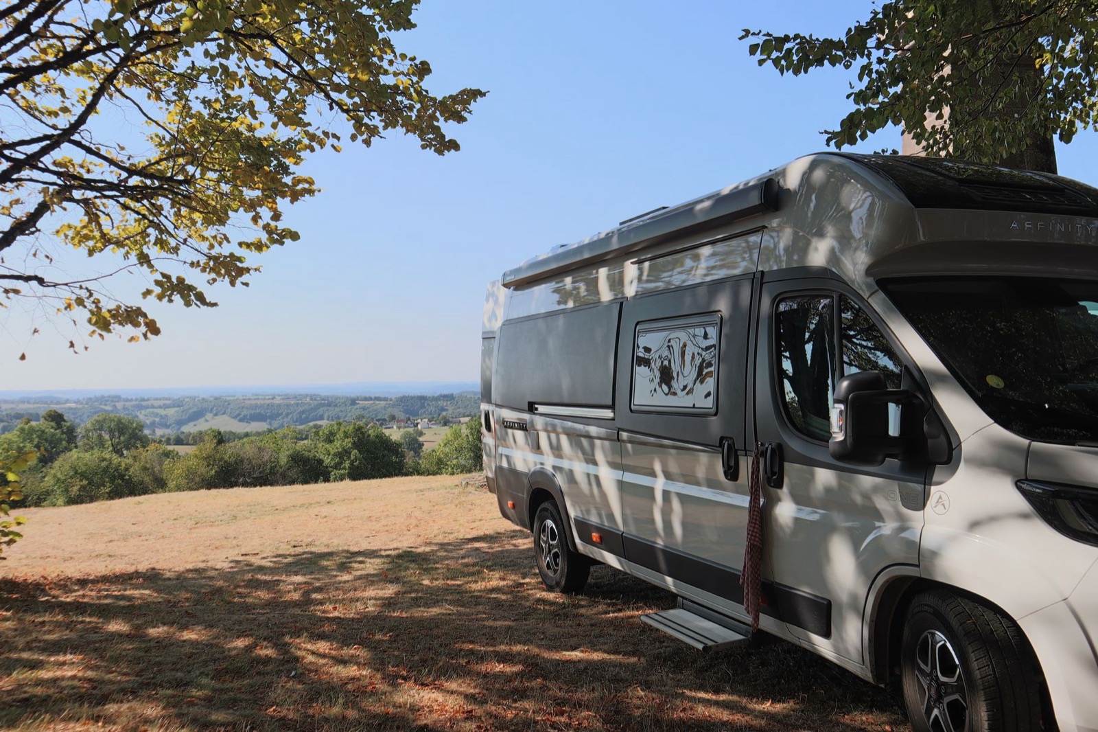 A camper van parked in tree shade on a grassy hilltop above rolling wooded country