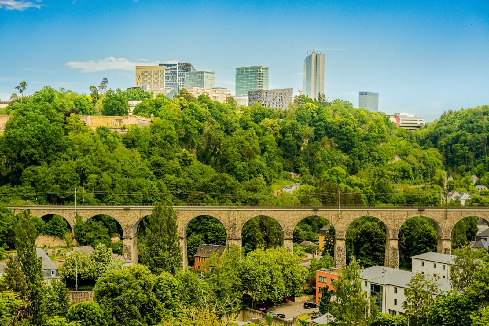 A stone railway viaduct over a green valley below the glass towers of Kirchberg in Luxembourg City