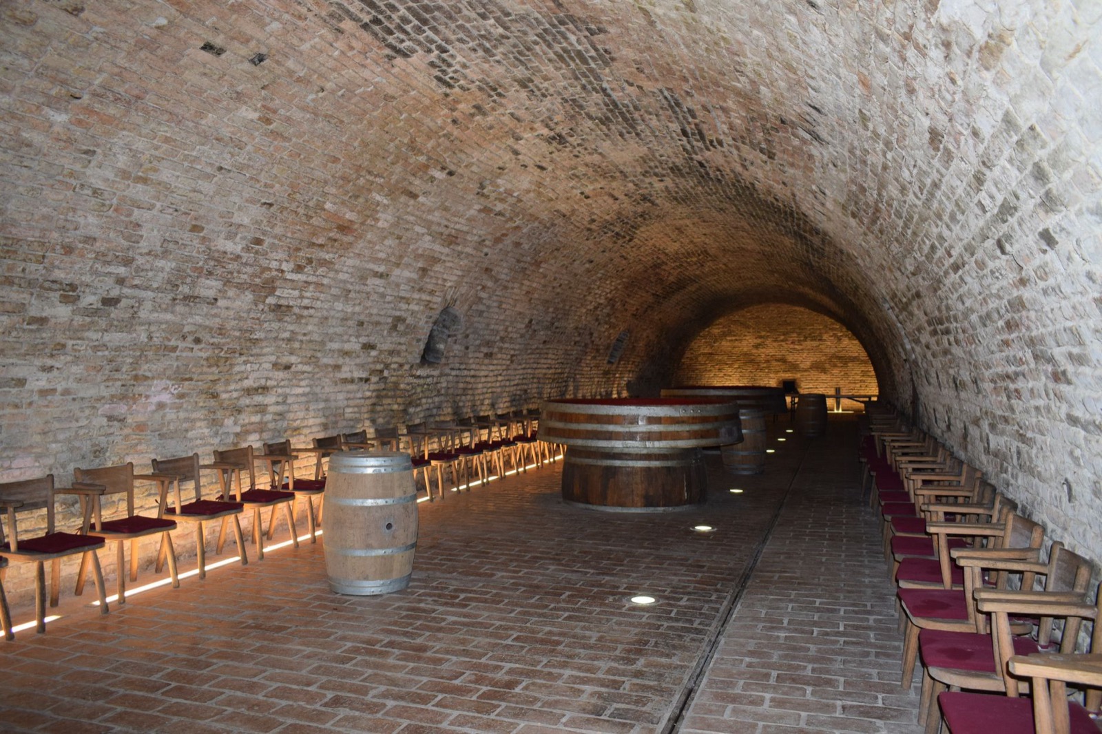 A brick barrel-vaulted cellar with wine casks and rows of chairs along the walls