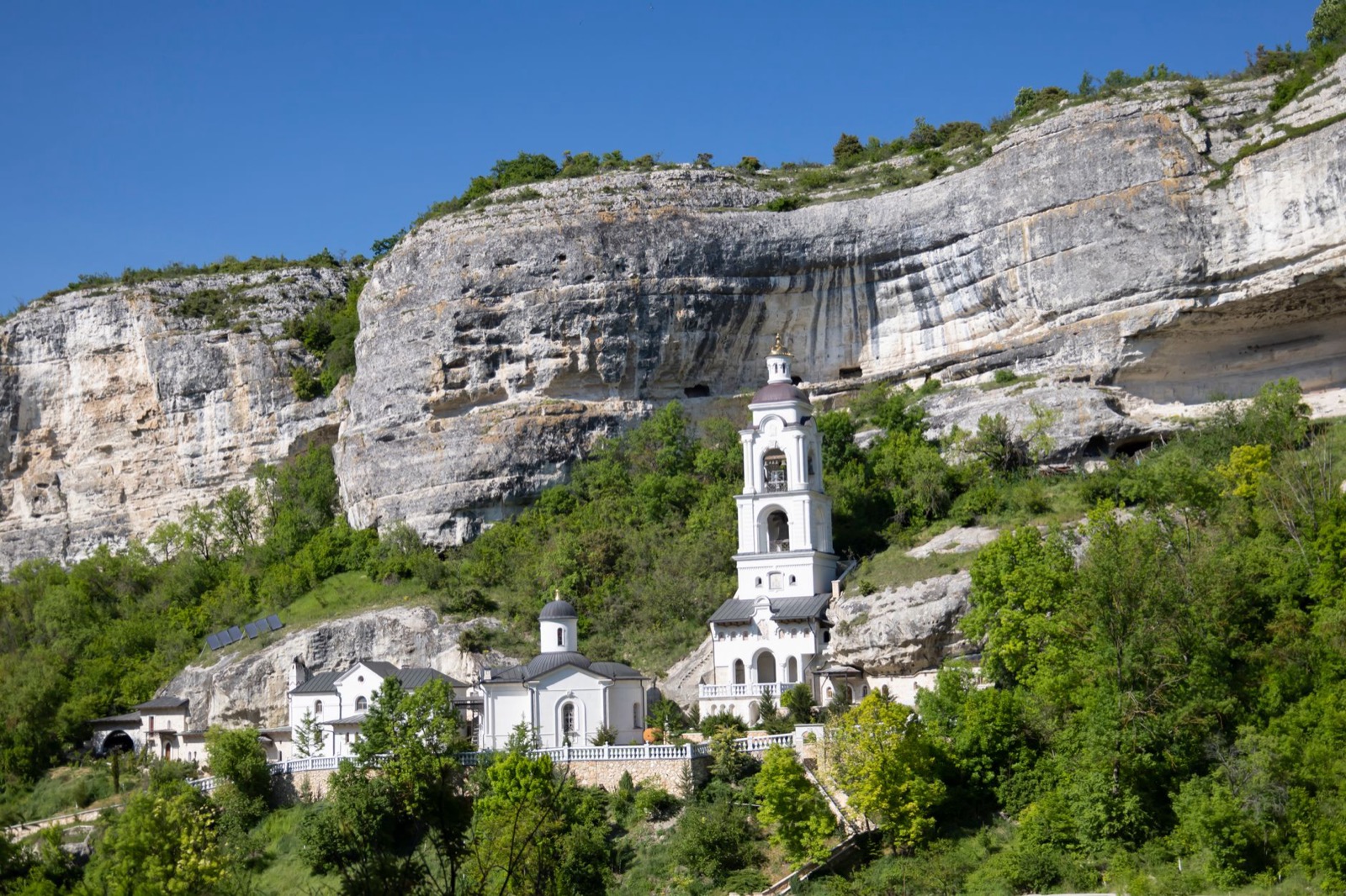 A white church and bell tower tucked beneath a huge limestone overhang above wooded slopes