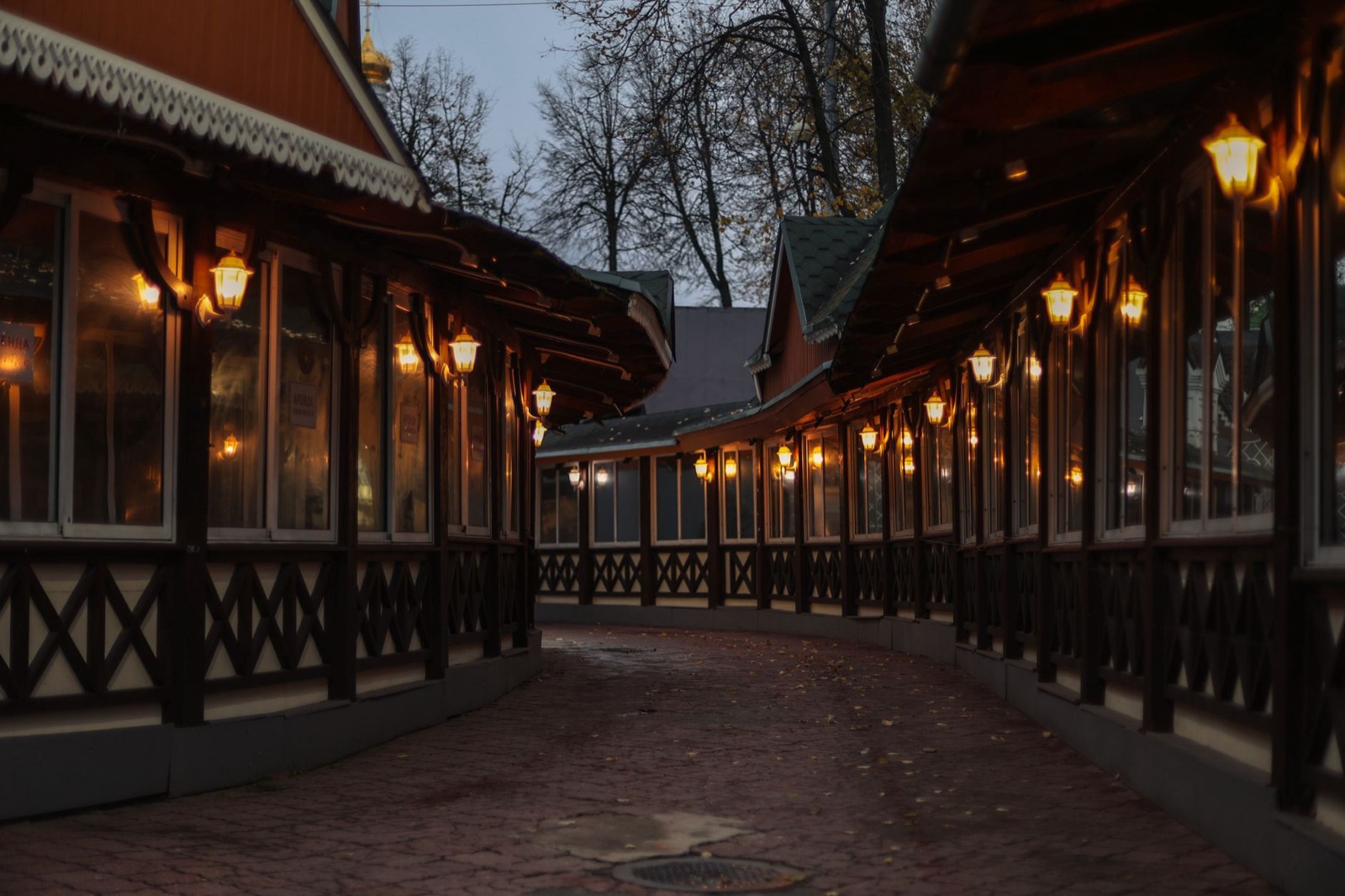 A covered wooden arcade hung with lanterns along an empty paved walkway at dusk