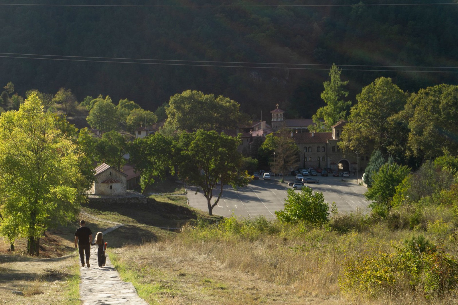 Two people walking a path down towards a monastery and its car park in a wooded valley at golden hour