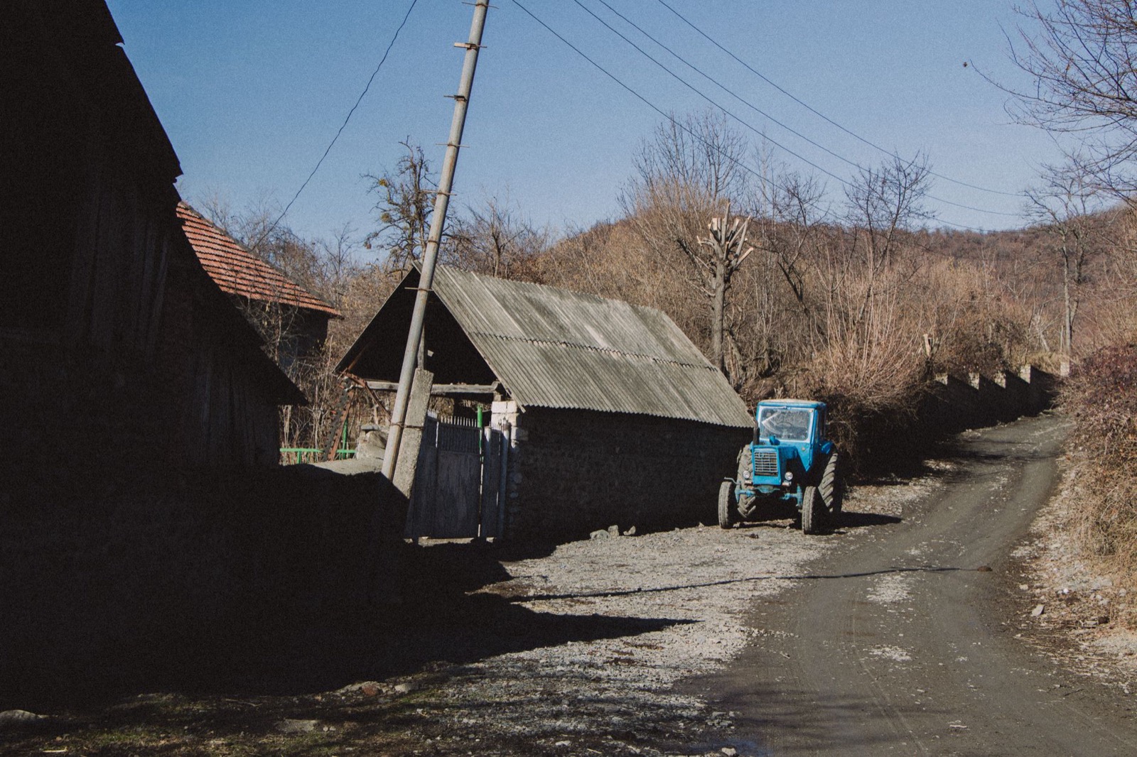 A blue tractor parked by weathered barns along an unpaved village lane in late winter