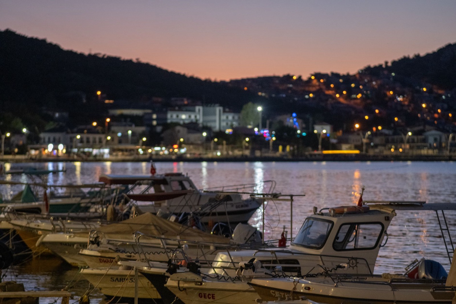 Small boats moored in a harbour at dusk below a hillside town lit up across the water
