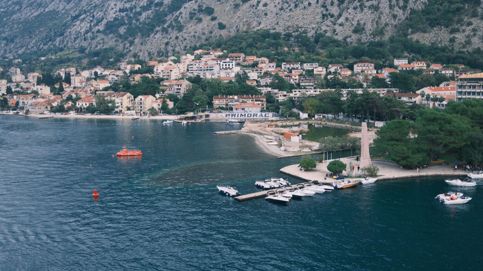 A village and its jetties on the shore of the Bay of Kotor seen from above, below a grey mountainside