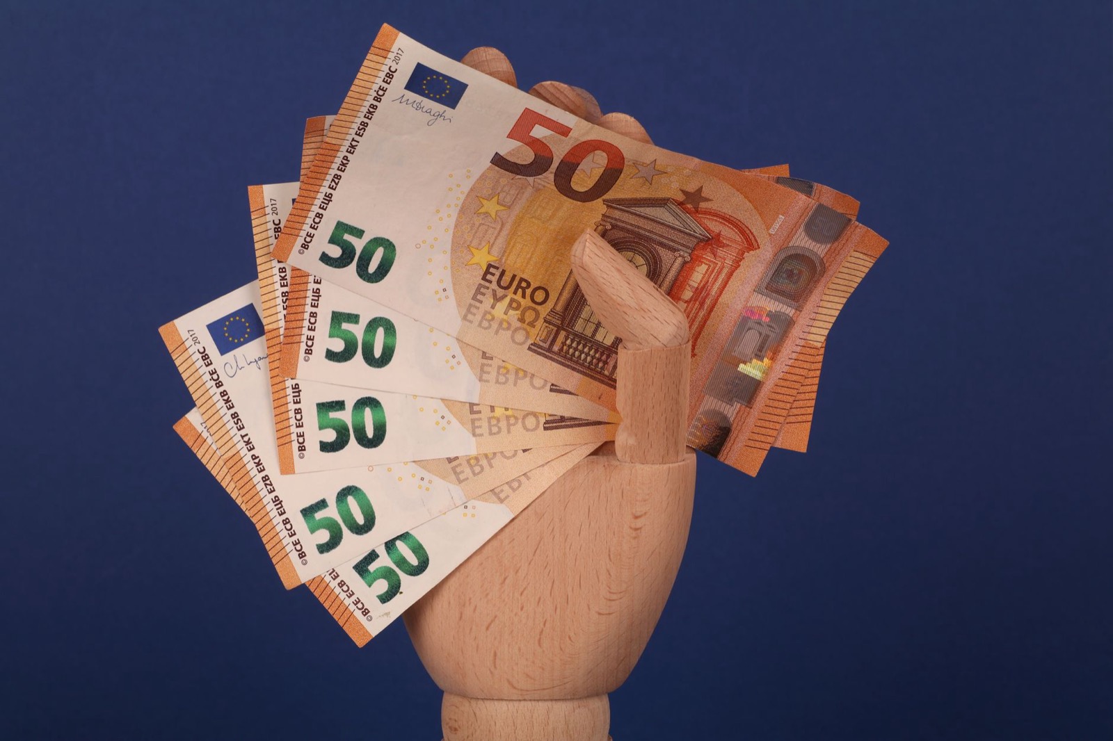 A wooden mannequin hand holding a fan of 50 euro notes against a dark blue background