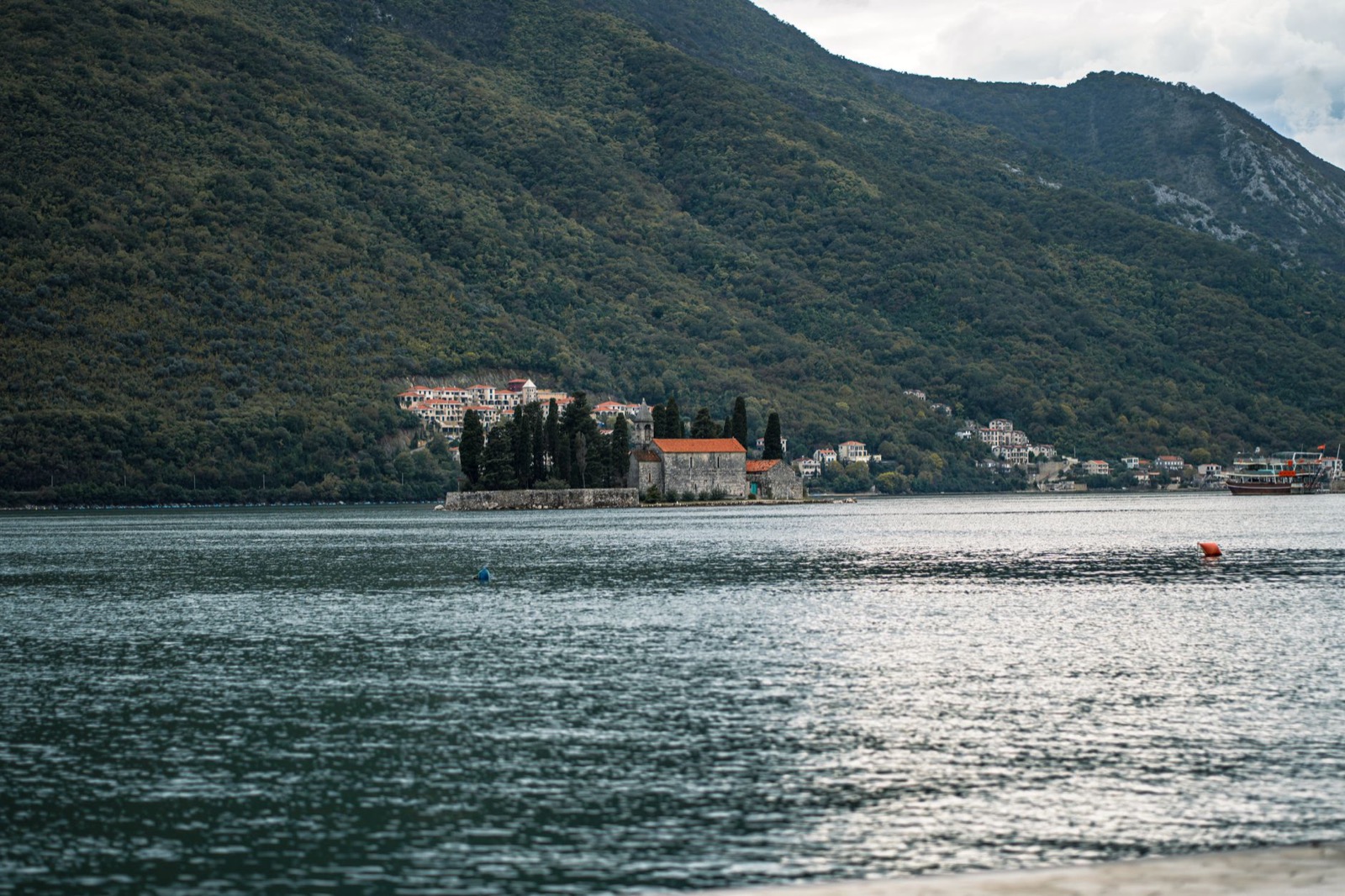 A small red-roofed church on a walled promontory jutting into the Bay of Kotor below wooded slopes
