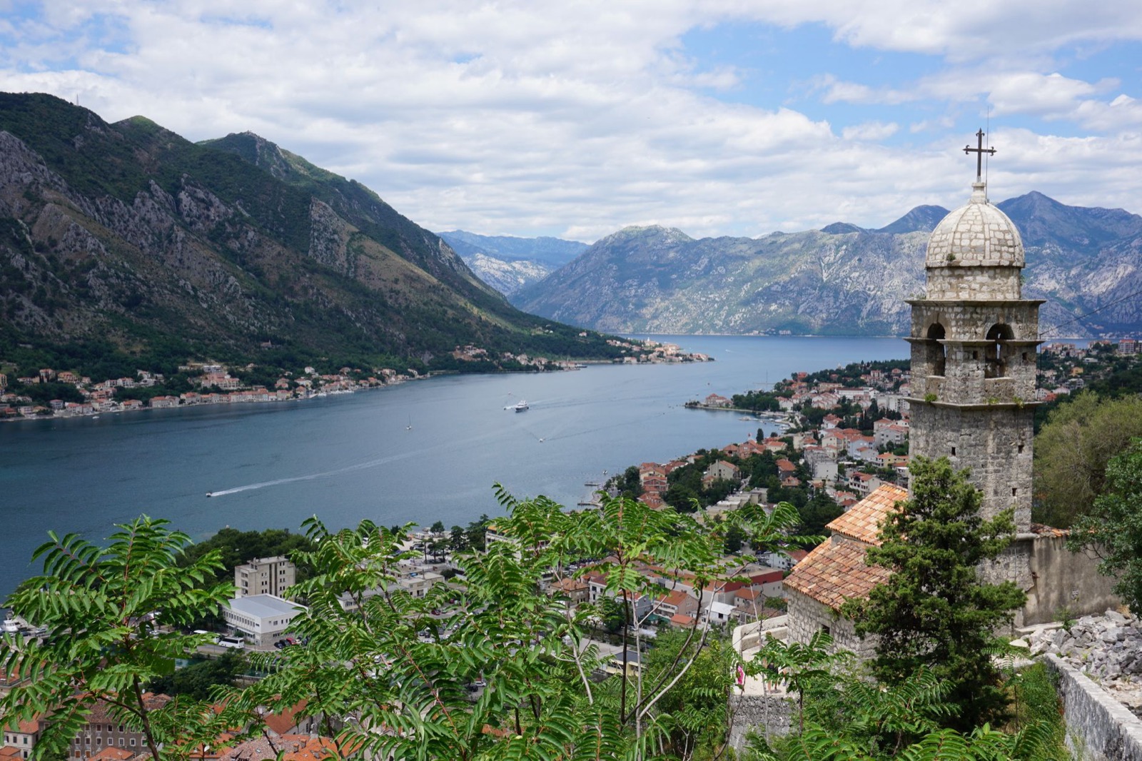 A stone bell tower on a steep green slope above the Bay of Kotor, with the town and the water below