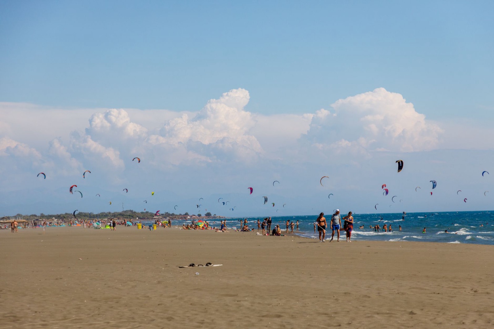 A long flat sandy beach crowded with bathers under dozens of kitesurfing kites