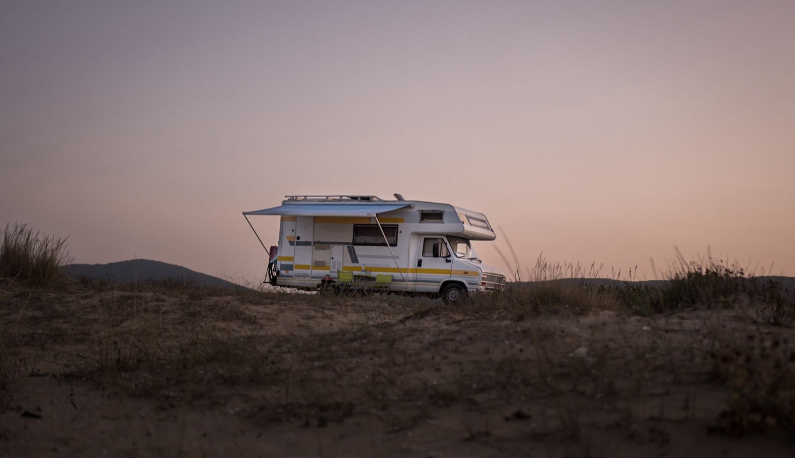 An older coachbuilt motorhome standing on dry scrubland at dusk with its awning part way out