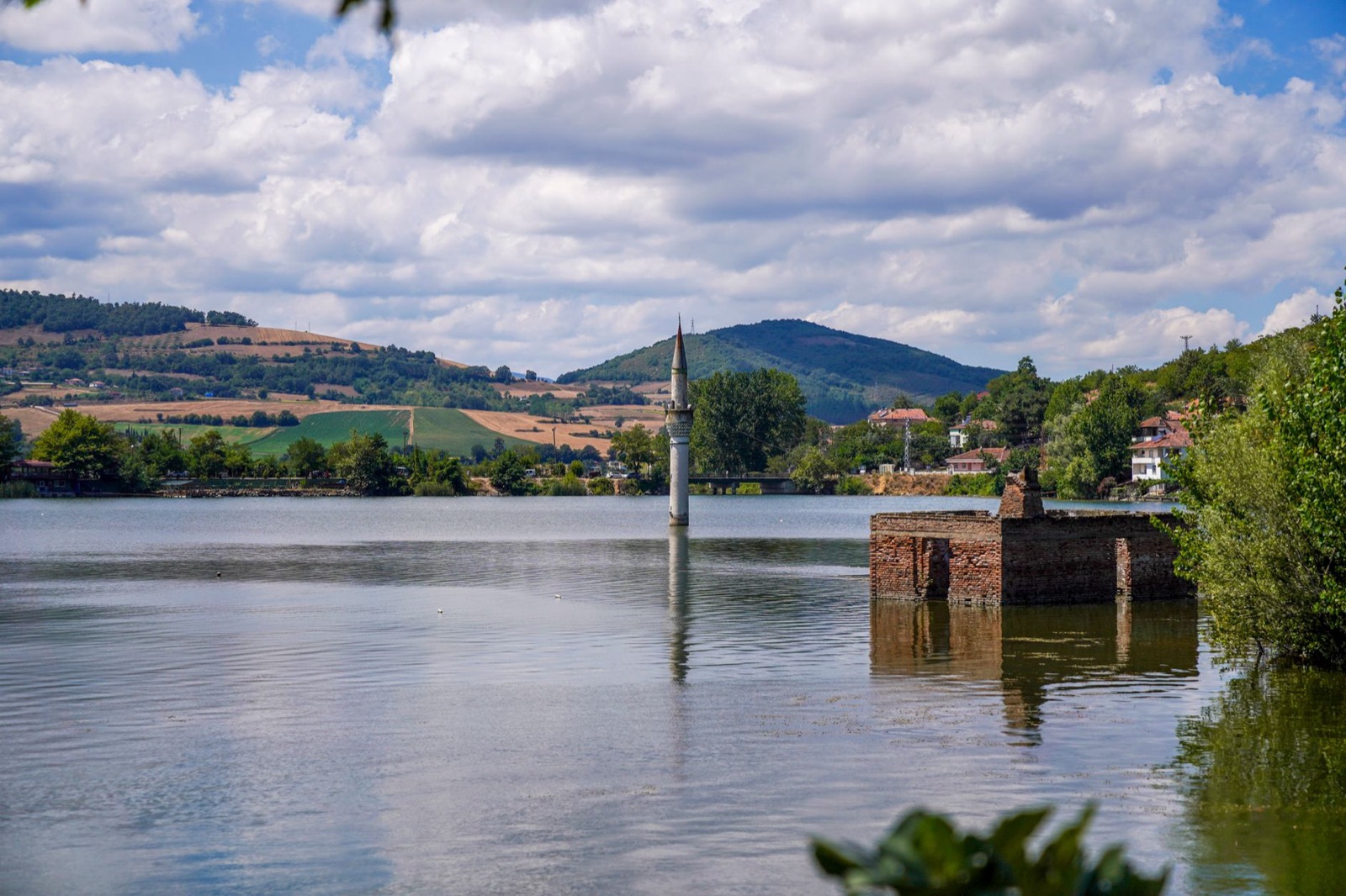 A half-submerged minaret and ruined brick walls standing in a reservoir below green hills