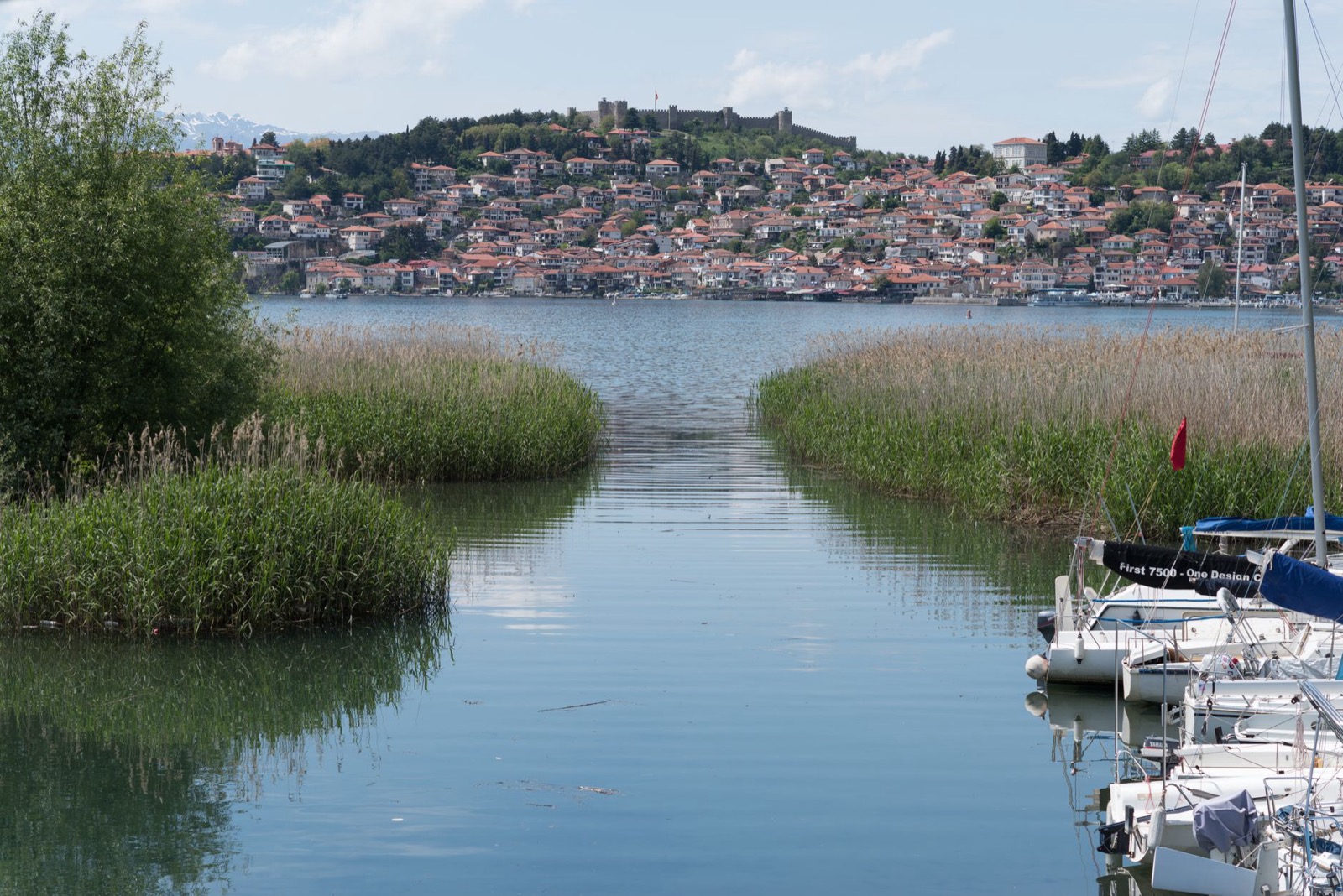 A reed-lined channel opening onto the lake, with Ohrid's terracotta roofs and hilltop fortress across the water
