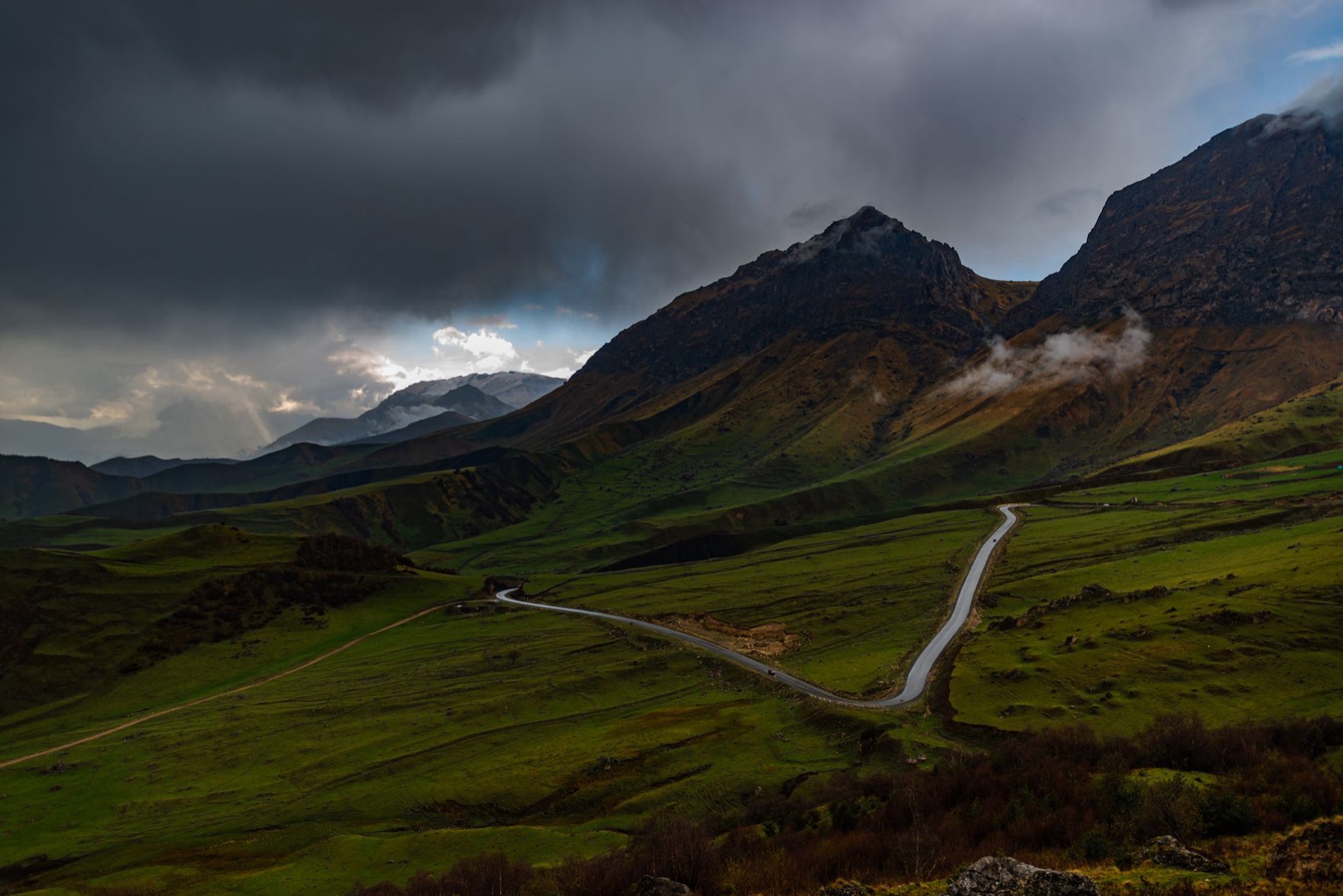 A road winding across a green highland valley below dark storm cloud