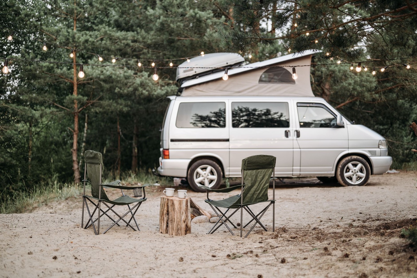 A camper van with its pop-top roof raised in a pine wood, with two camping chairs and festoon lights strung above