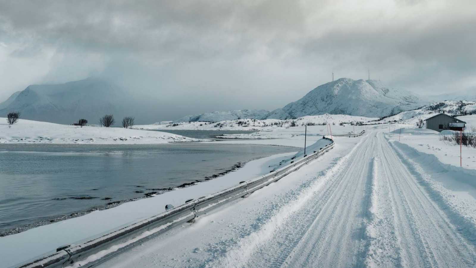 A snow-covered road running beside open water below low mountains in winter