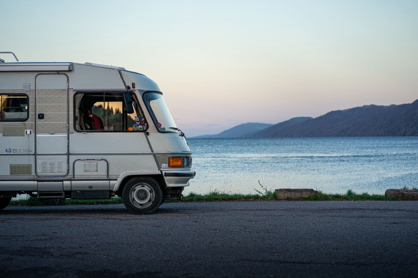 A white motorhome parked at the edge of a road beside calm water at dusk