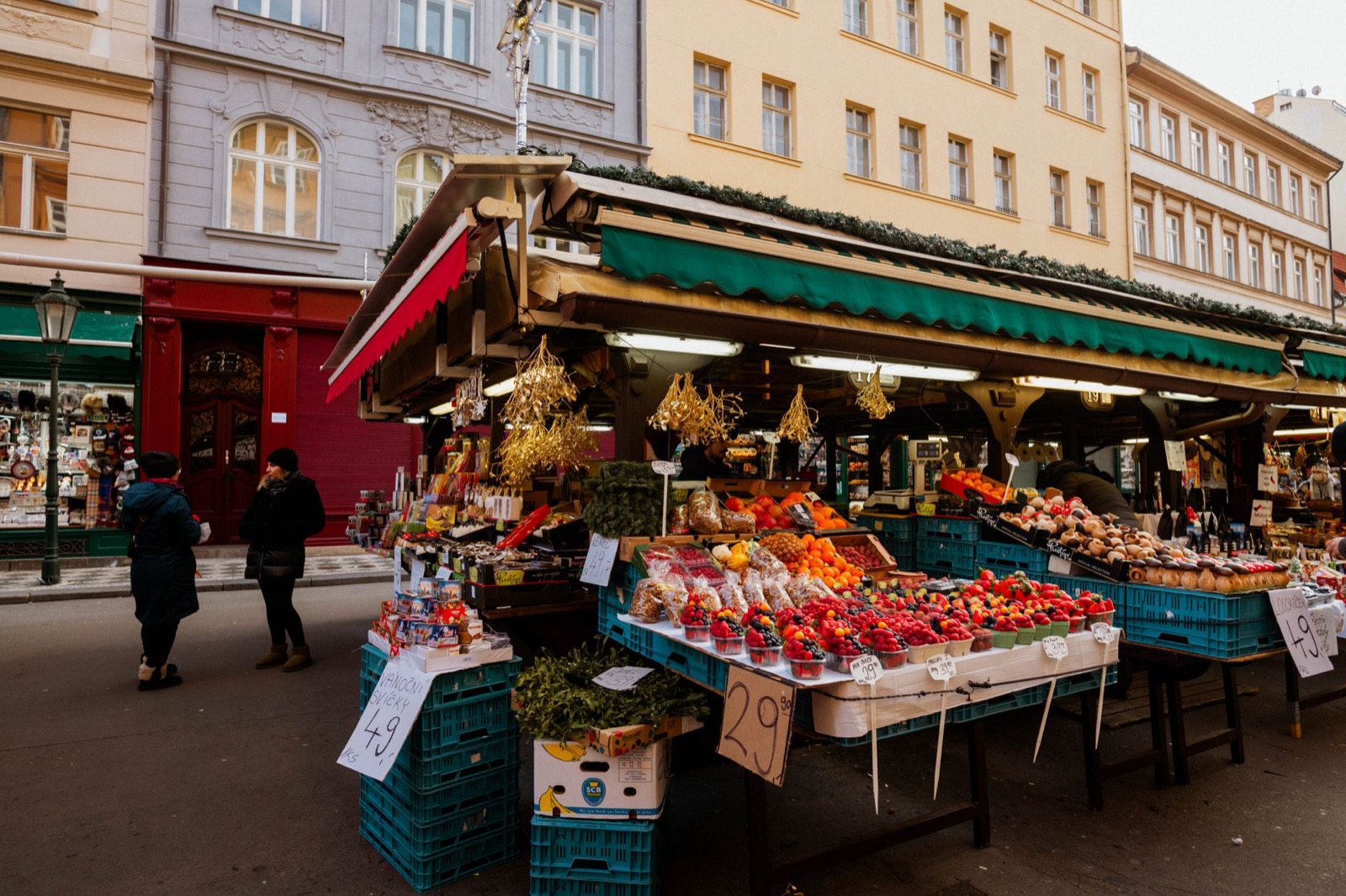 Fruit and vegetable stalls under a green awning on a city street, with handwritten price cards