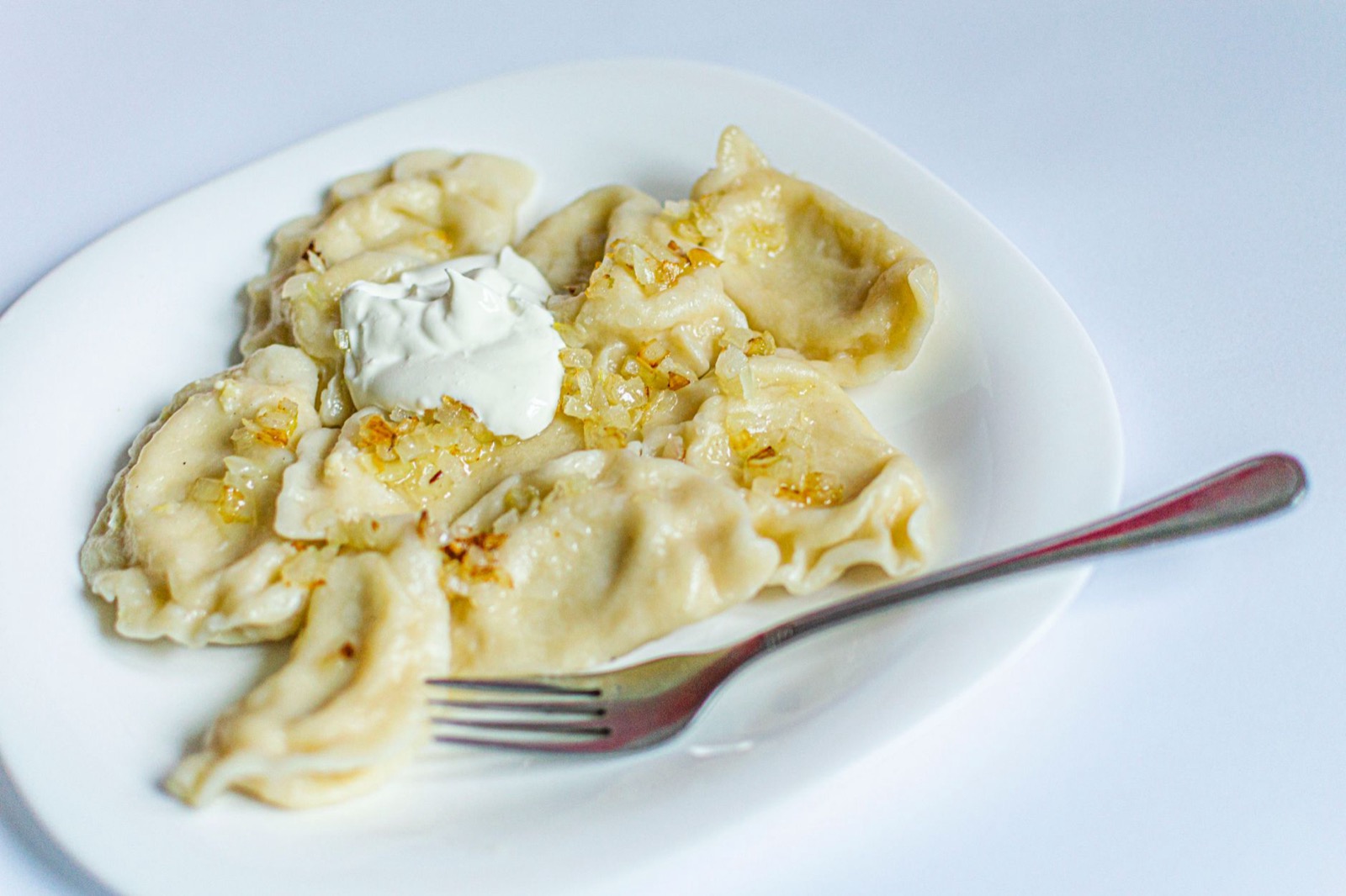 A plate of boiled pierogi topped with fried onion and a spoonful of sour cream