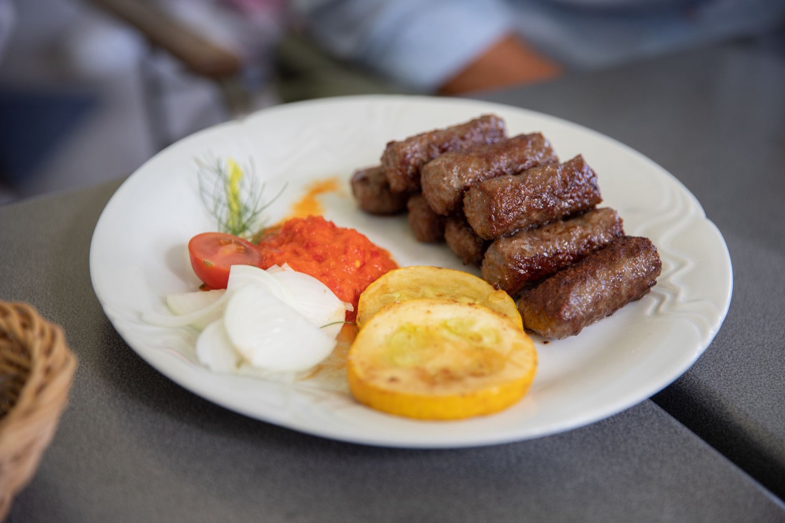 Grilled minced meat rolls on a white plate with ajvar, raw onion and tomato