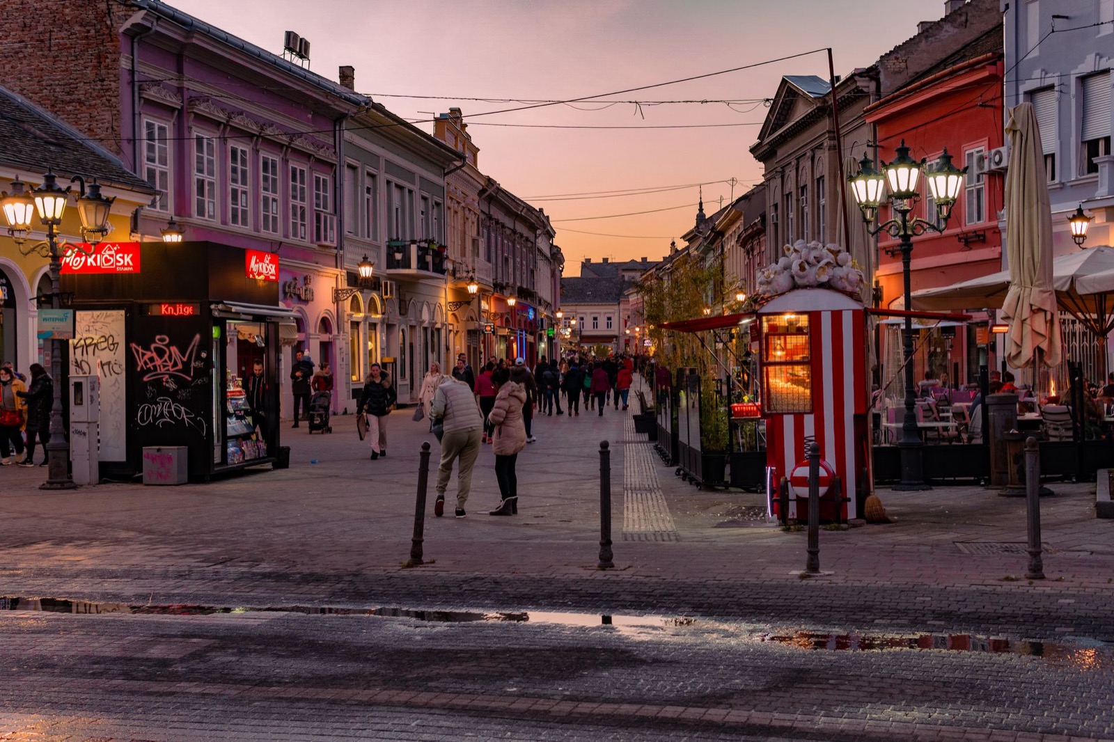 A lit pedestrian street at dusk with people walking past kiosks and cafe terraces
