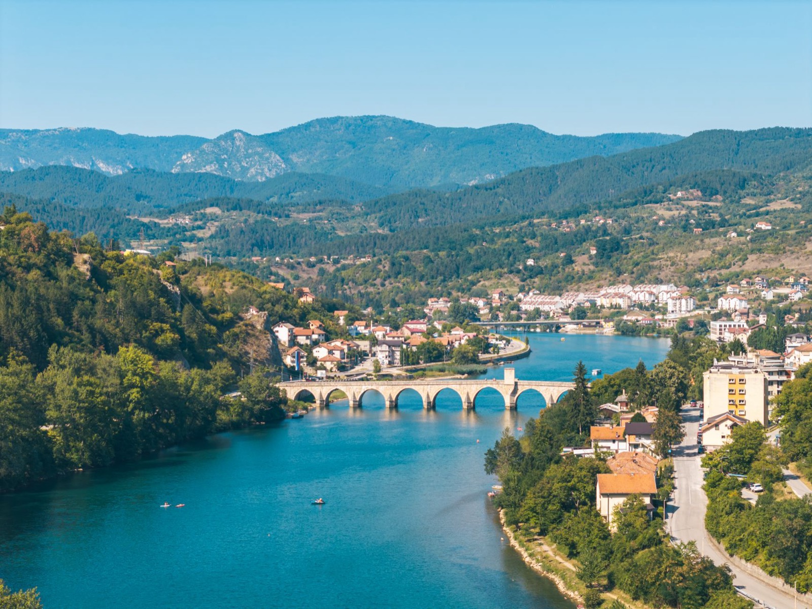A turquoise river curving past a town and a long arched bridge below wooded hills