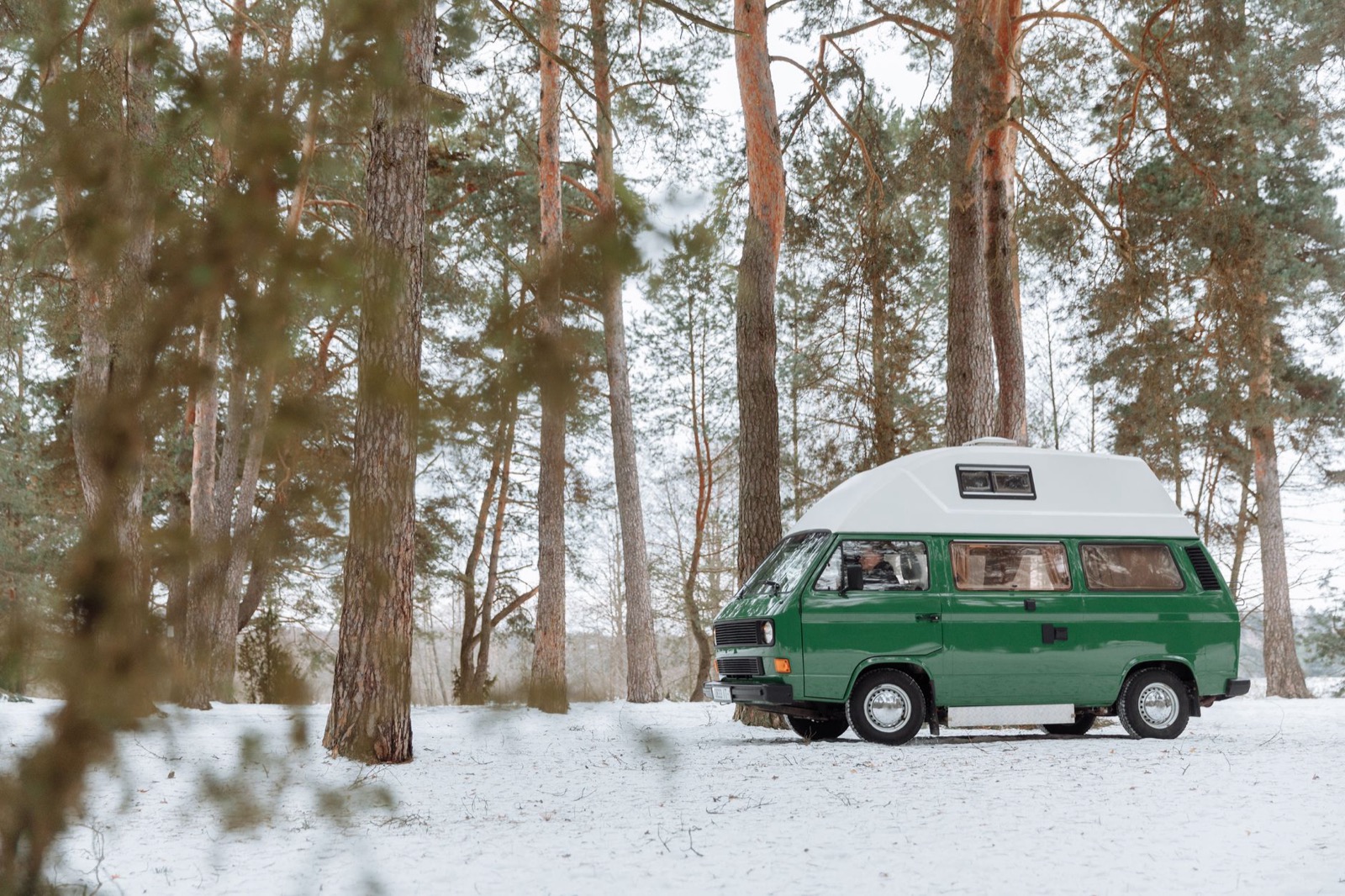 A green camper van with a high roof parked among pines in a snowy forest