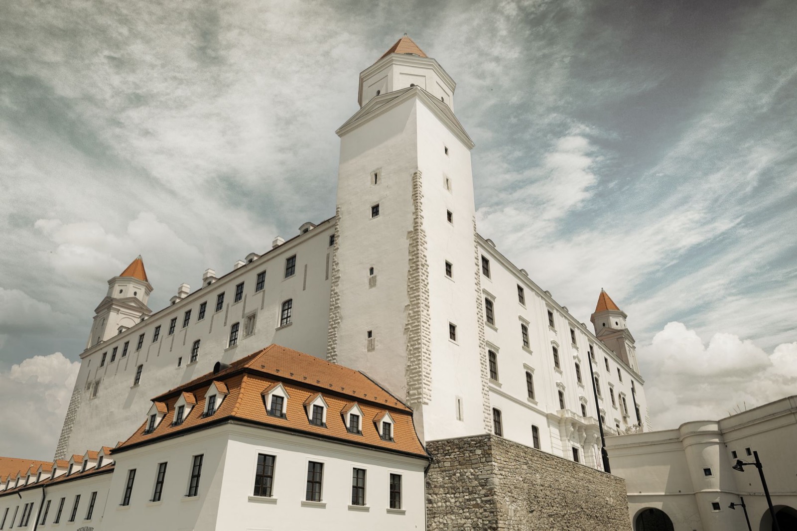 The white walls and corner towers of Bratislava castle seen from below against a streaked sky