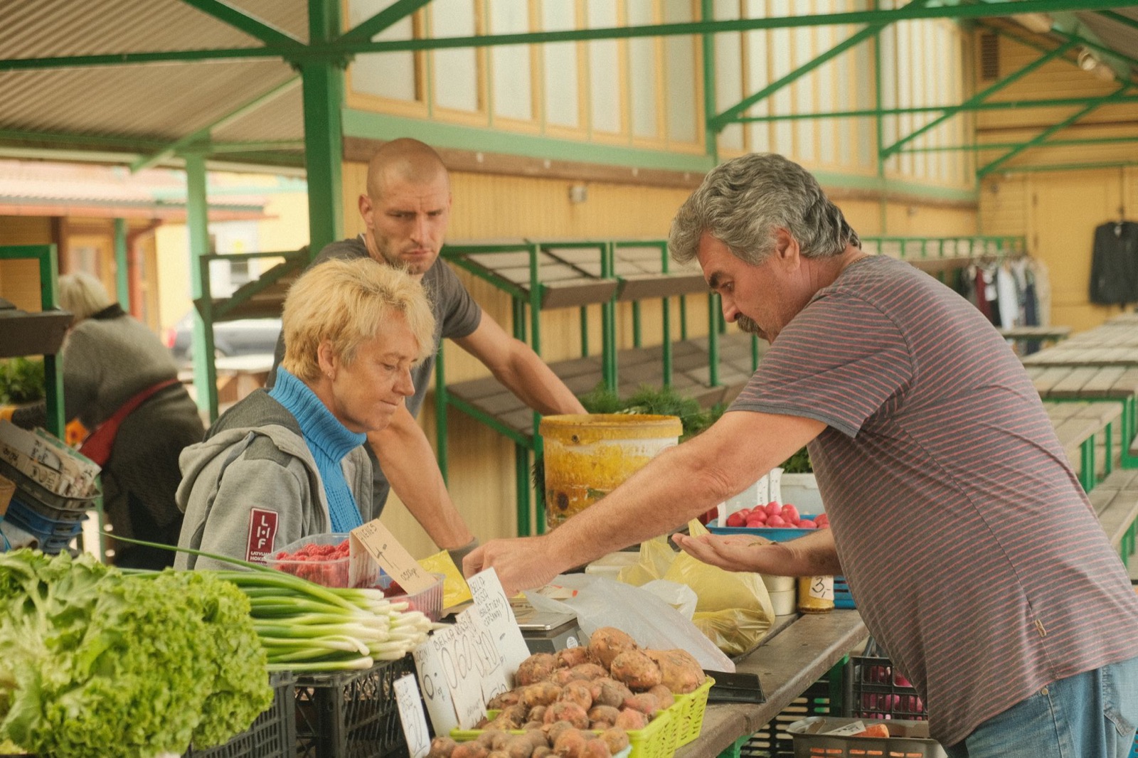 A trader handing change across a market counter of potatoes, spring onions and lettuce under a green steel canopy