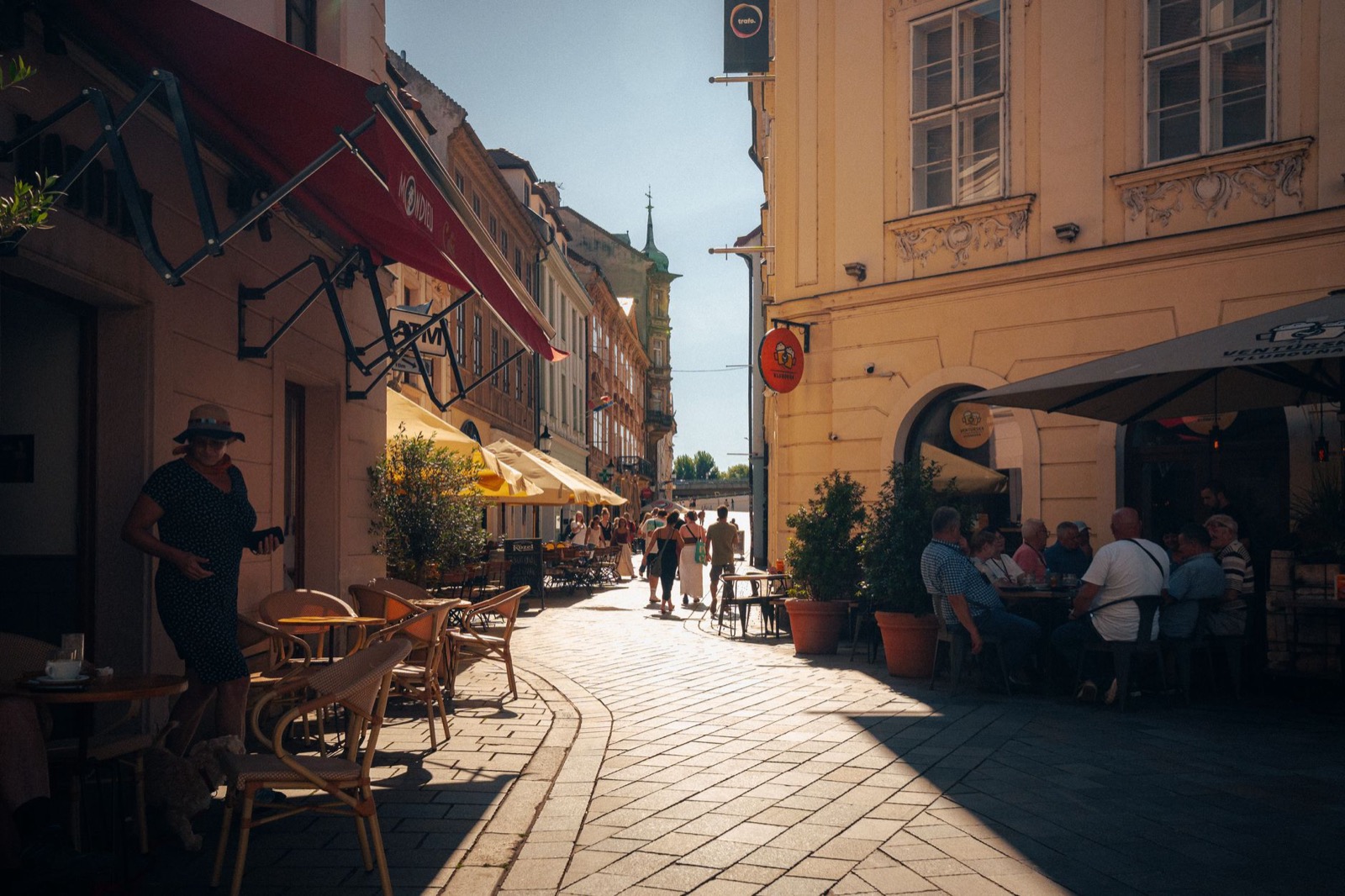 A sunlit old town lane with café awnings, outdoor tables and people walking between painted facades