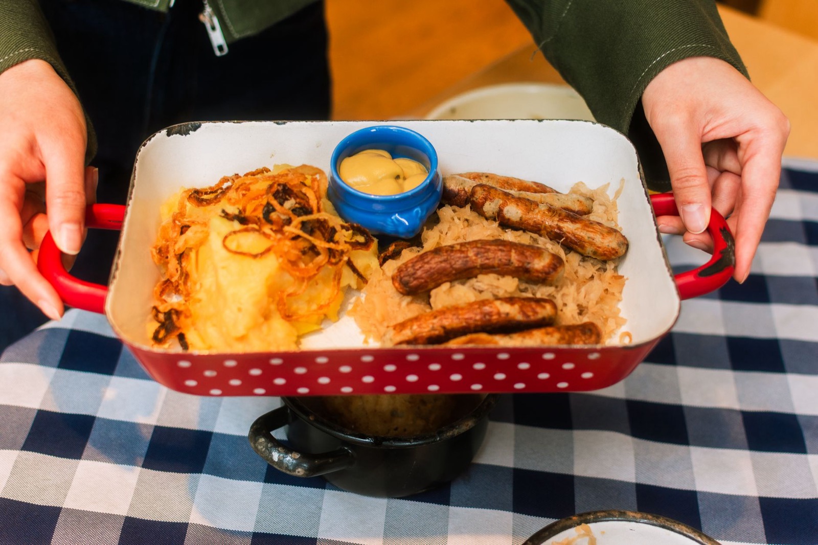 Grilled sausages with sauerkraut, mashed potato and mustard in a red enamel pan held over a checked cloth