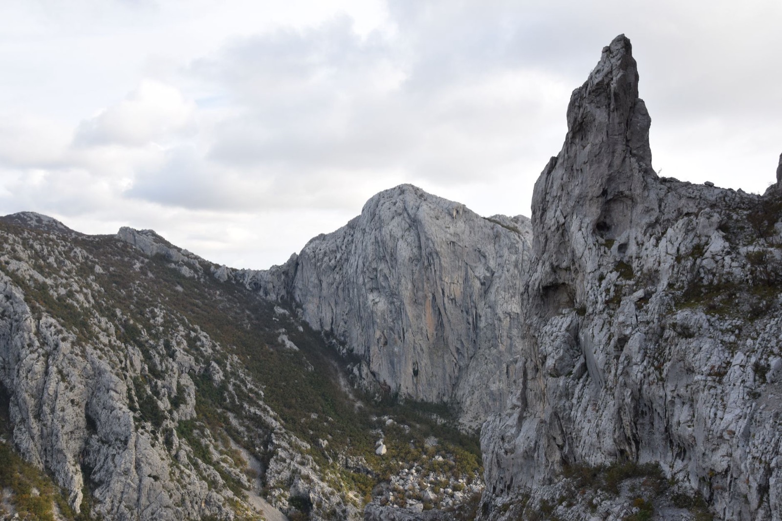 Bare limestone crags and a tall rock pinnacle above a scrubby valley under grey cloud
