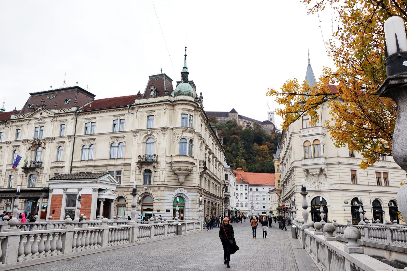 The balustraded Triple Bridge in Ljubljana with pale facades around it and the castle on its wooded hill behind