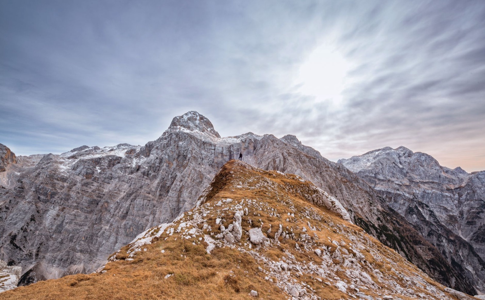 A single walker on a grassy ridge below jagged limestone peaks under a pale sky