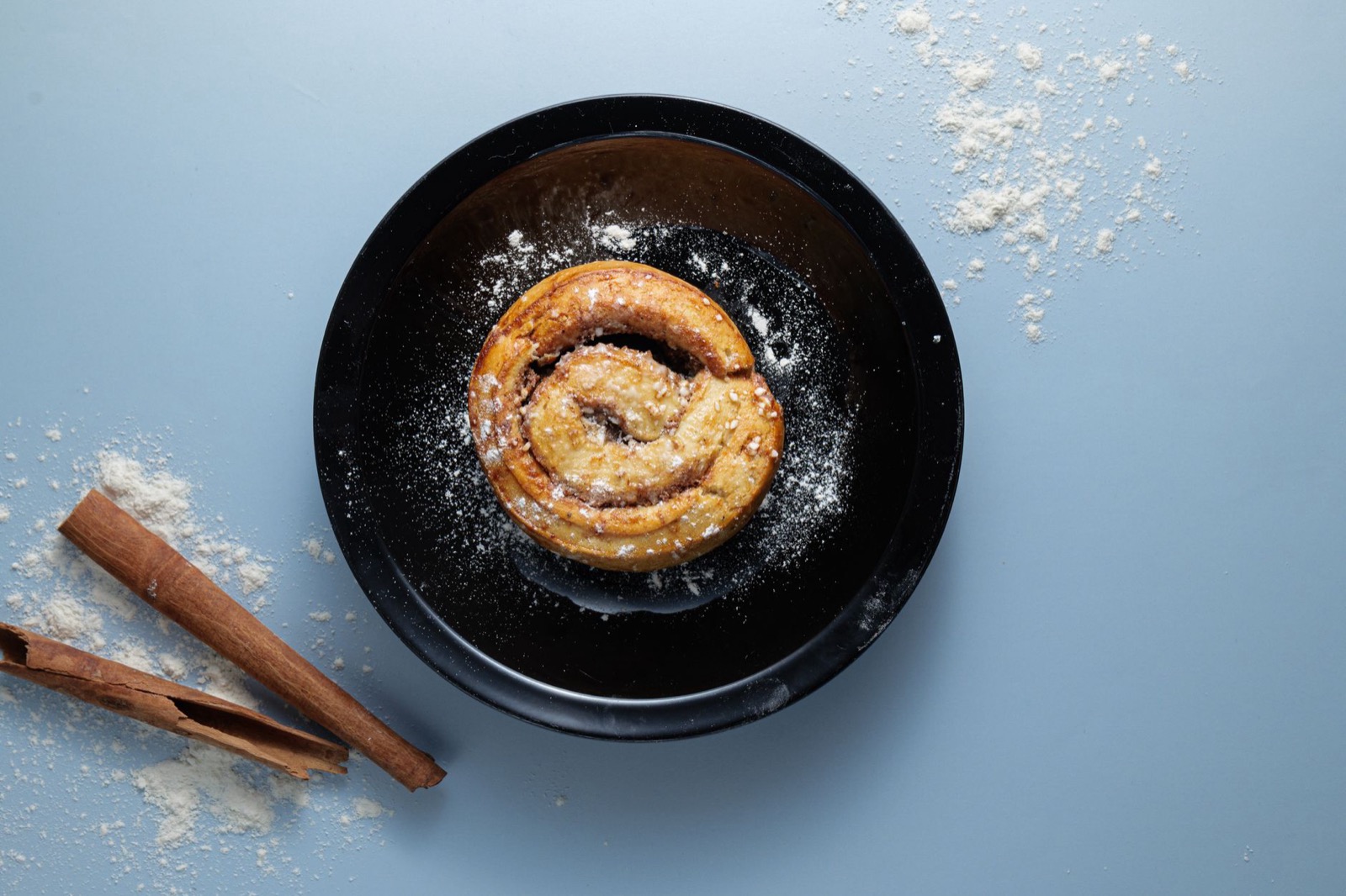 A cinnamon bun dusted with sugar on a black plate beside cinnamon sticks