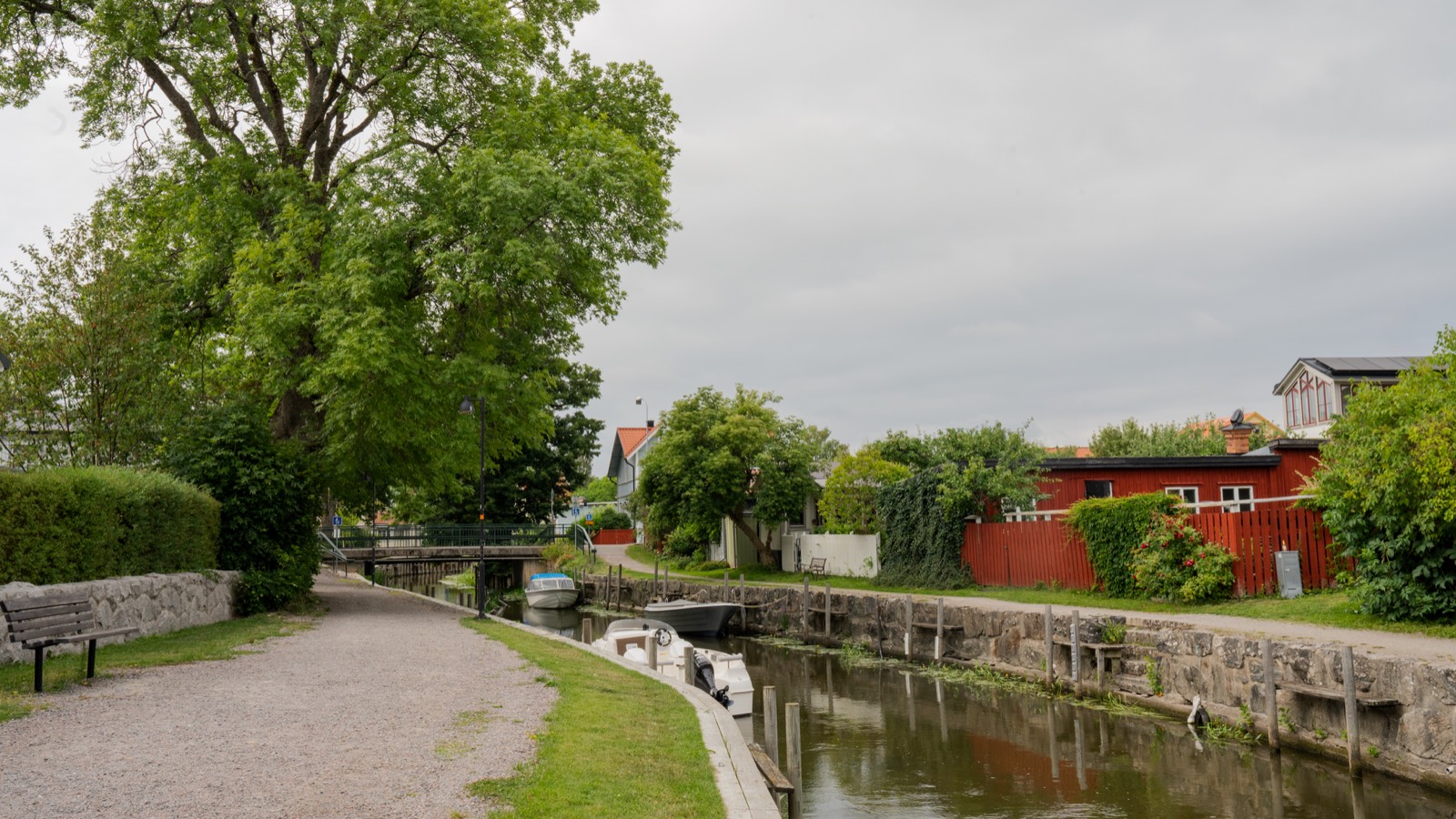A gravel path beside a narrow canal, with red wooden buildings and trees along the far bank